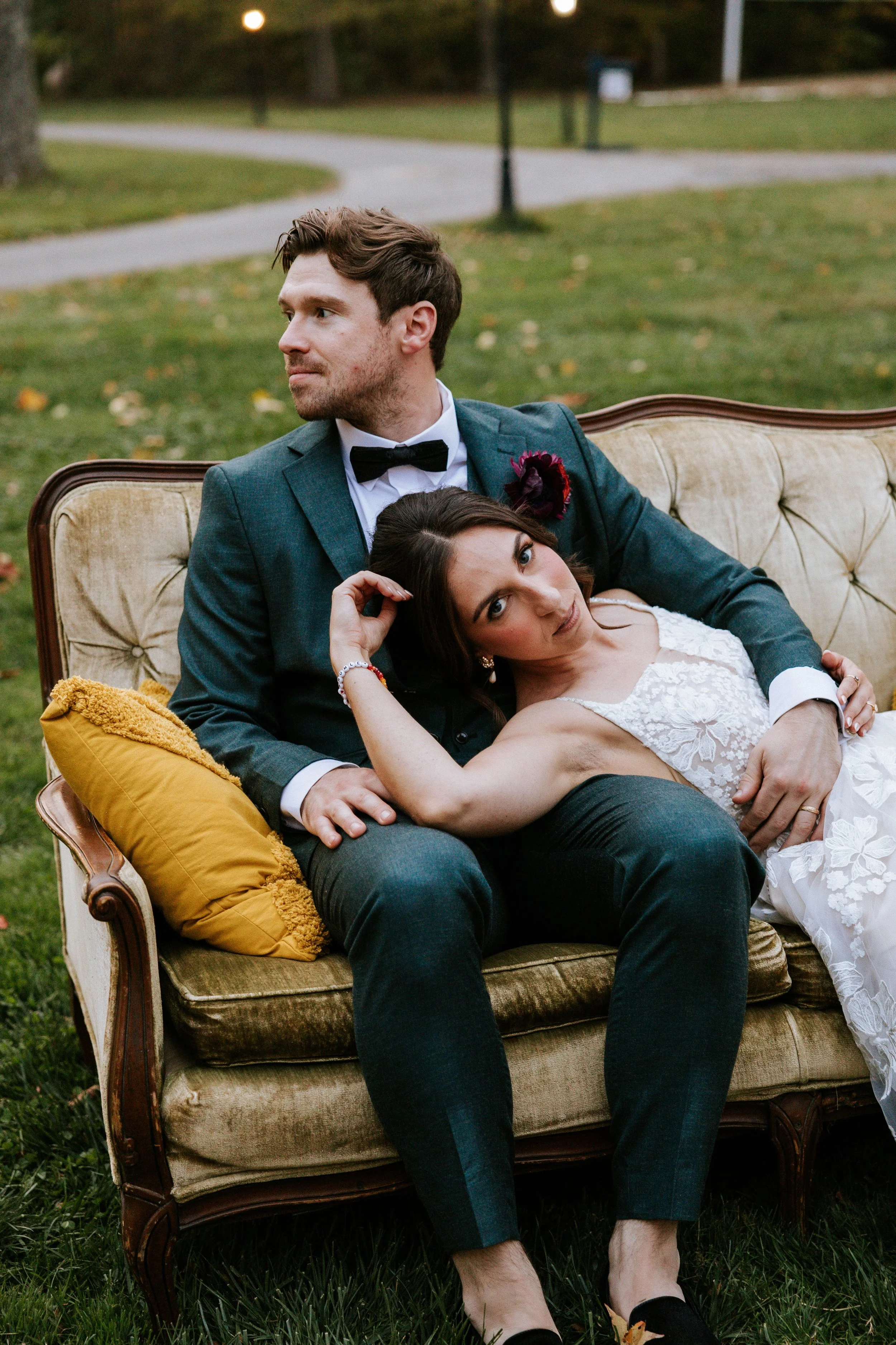 A man and woman sitting on a vintage sofa outdoors. The man is wearing a dark suit with a bow tie, and the woman is in a white lace dress. They are relaxing on the sofa on green grass, with the woman resting her head on the man's chest and looking in