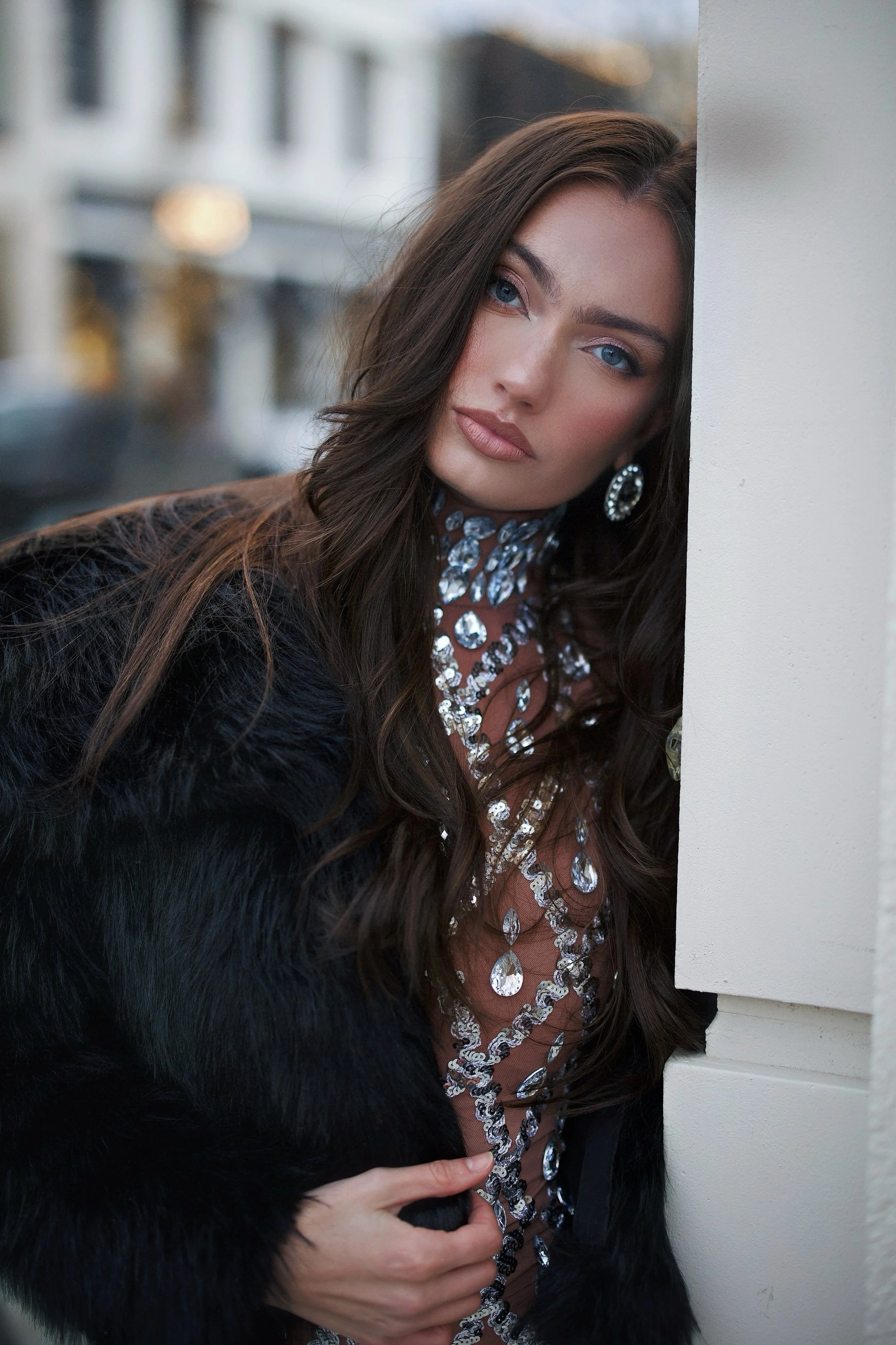 A young woman with long brown hair, blue eyes, and wearing large earrings, a sparkly necklace, a beige top, and a black fur coat, is standing against a white wall and looking at the camera.