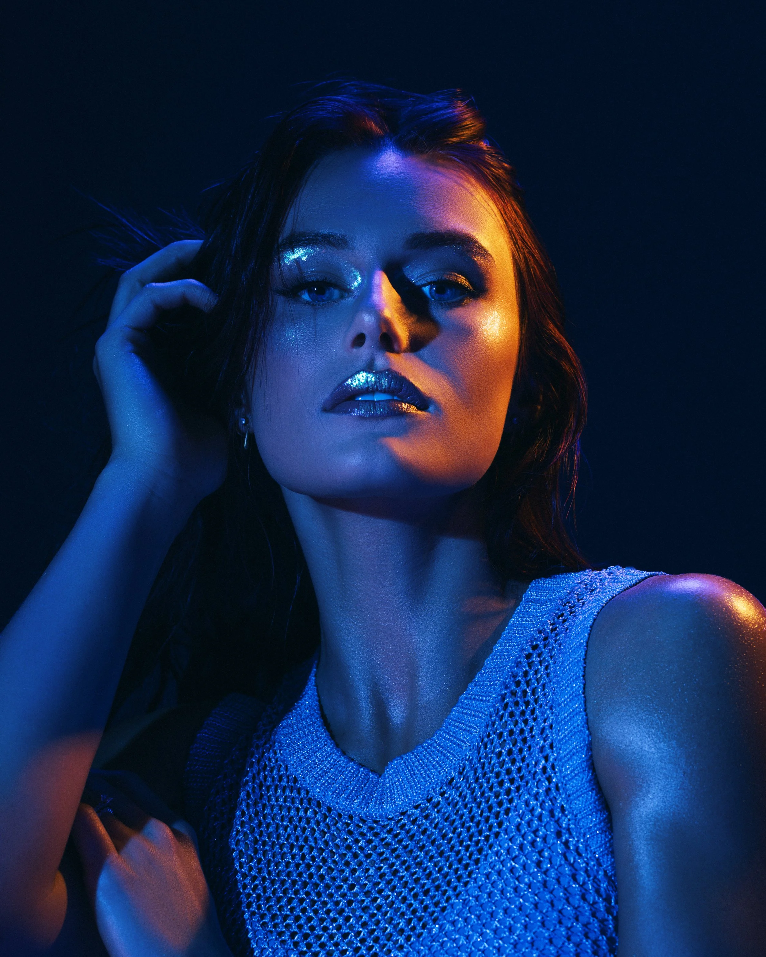 A woman with dark hair and blue eyes posing in low, colorful lighting, wearing a sleeveless, textured dress with metallic or sequined fabric.
