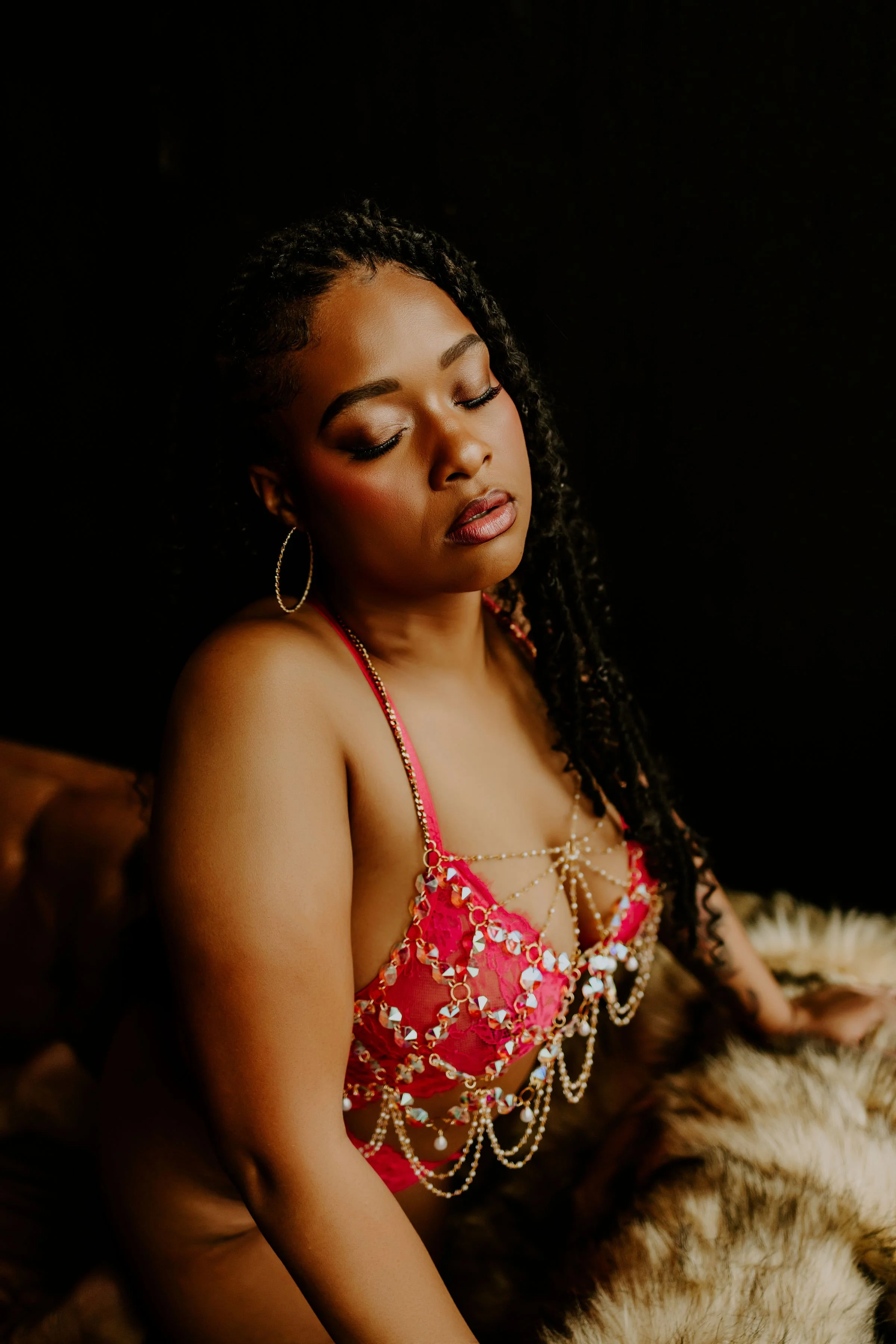A woman with closed eyes, wearing makeup, large hoop earrings, and a red, bejeweled, strappy top, sitting on a tiger-print fur surface against a black background.