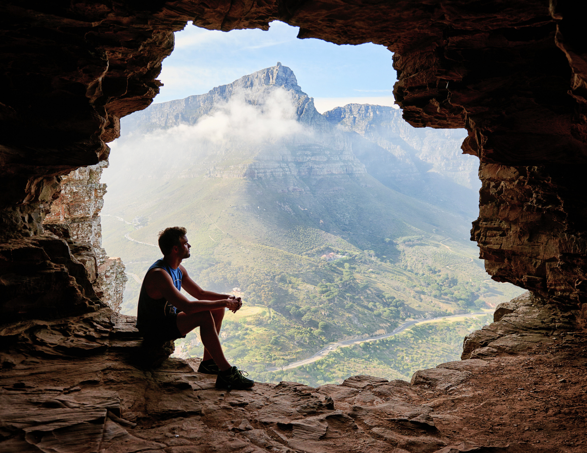 an image of a male sitting in a cave with the sun shining on a mountain he overlooks