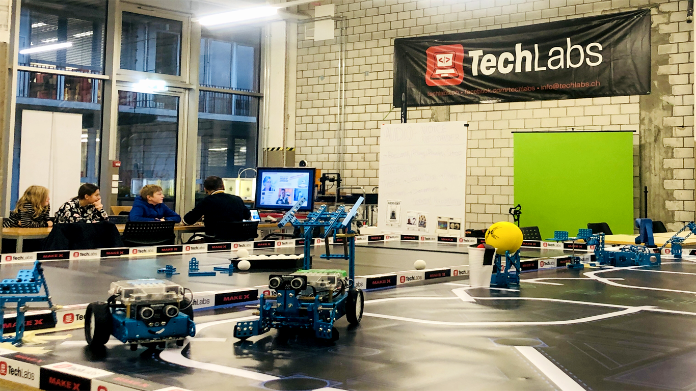 A robotics lab with technology equipment and a table where people are seated, discussing. A large banner reads 'TechLabs.' Several robotic components and a monitor with a video call are visible.