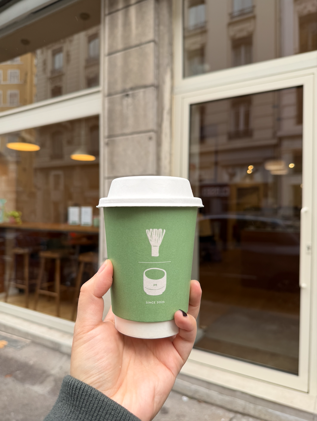 Une main tient une tasse à emporter en papier vert avec un couvercle blanc devant une vitrine de café