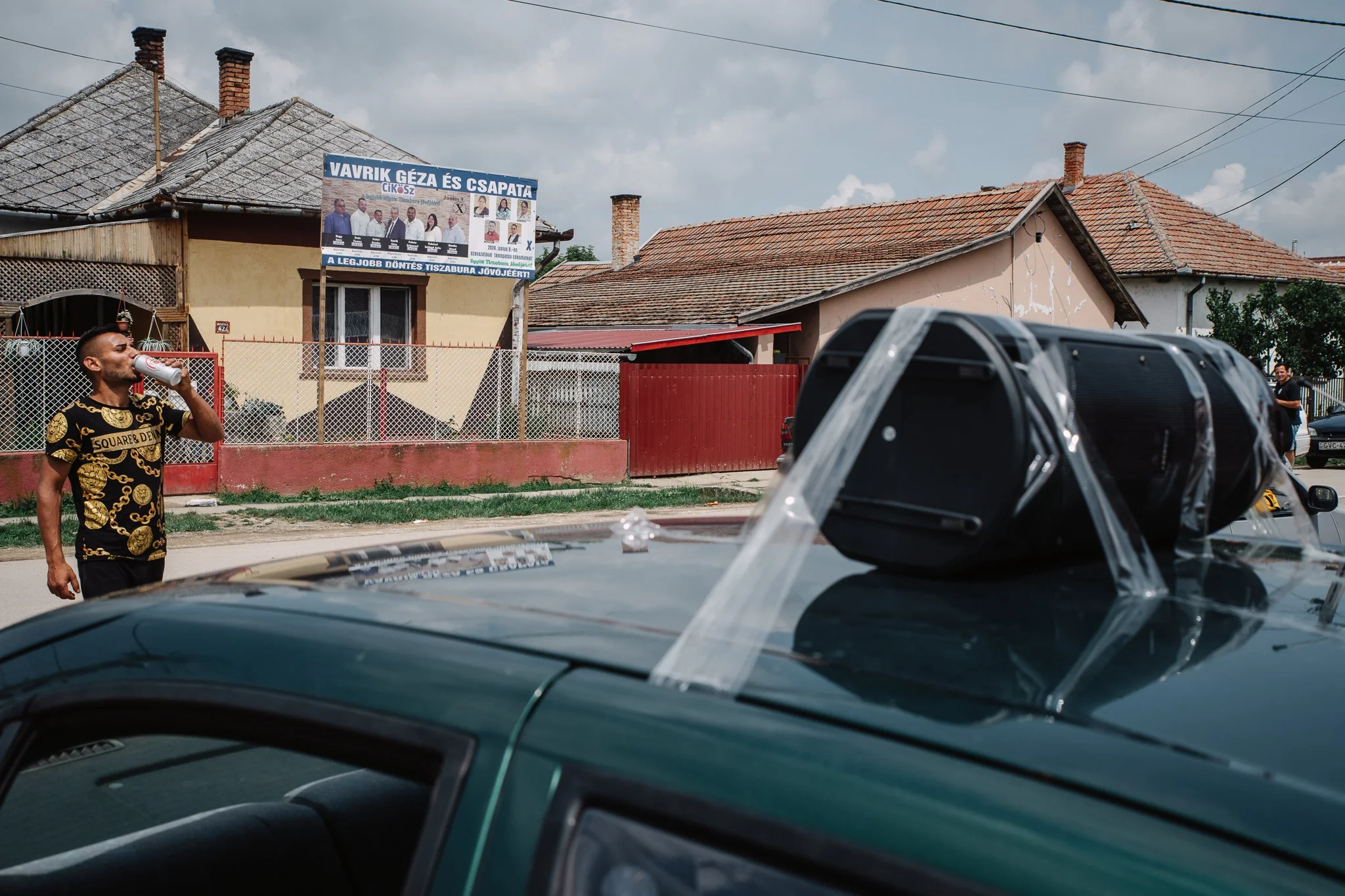 A man is drinking a beer on the side of the main road on the day of the elections. A huge Bluetooth speaker is mounted on the roof of a car. The car is used for mobilisation by the local CiKöSz team, whose poster can be seen in the background.
