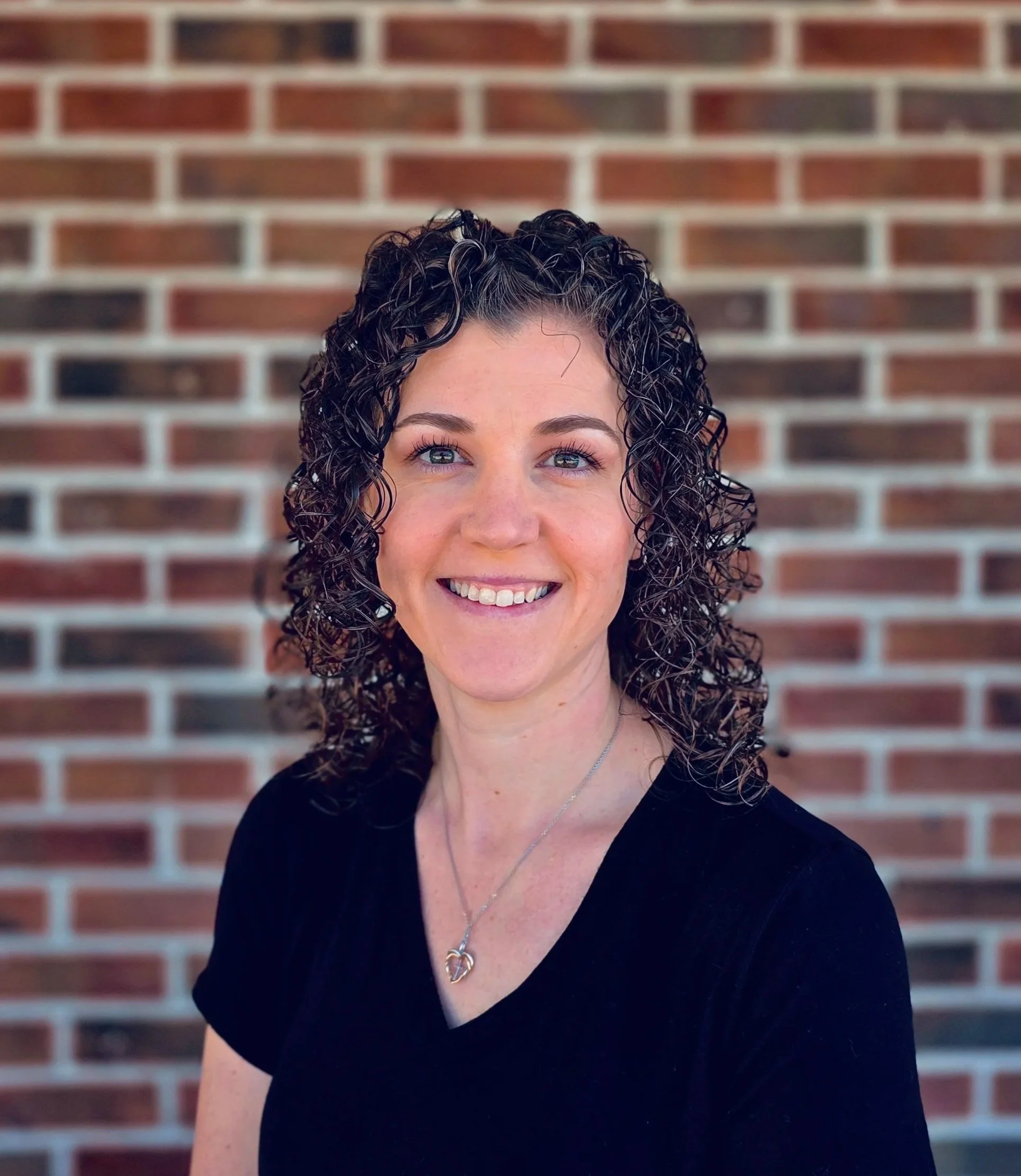 A smiling woman with long, curly brown hair standing in front of a brick wall.