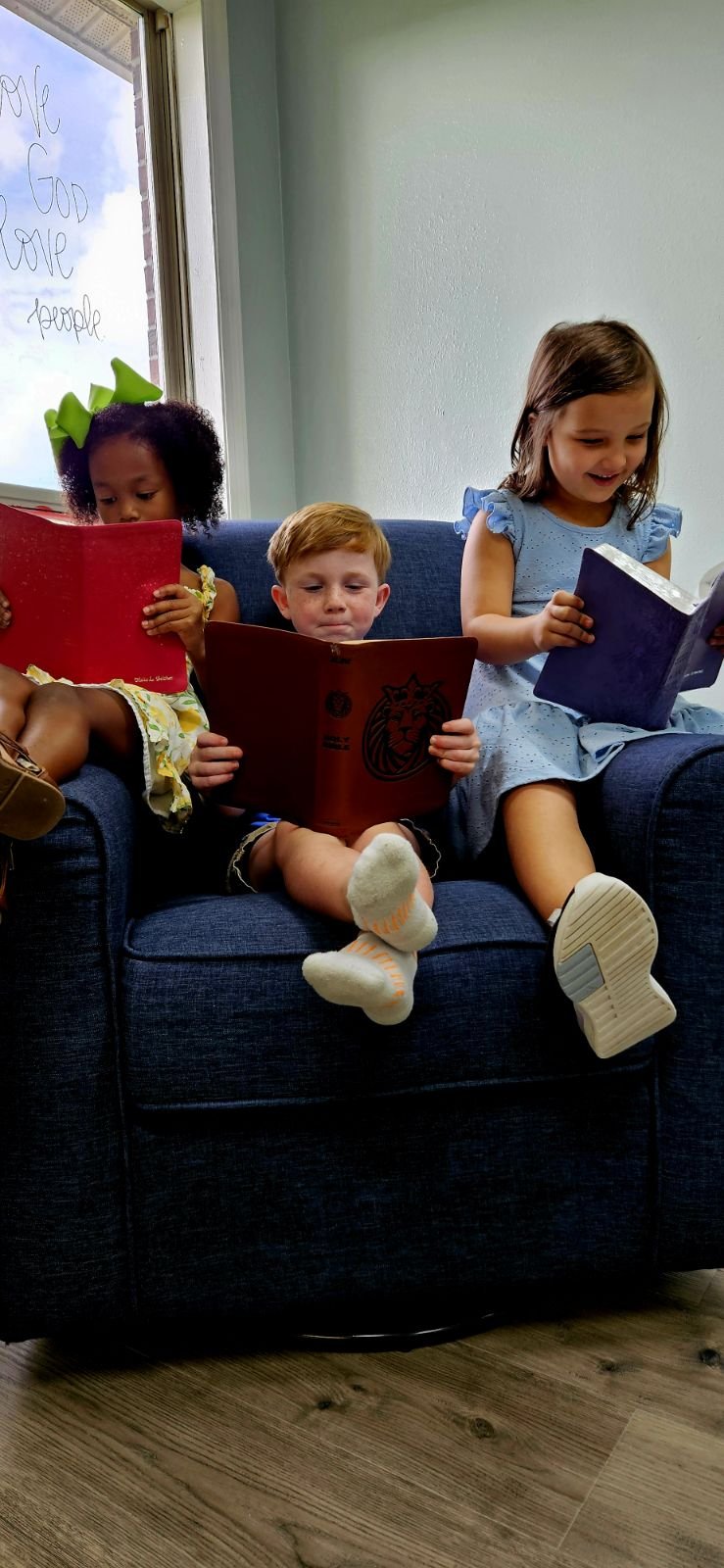 Three young children sitting on a blue couch, reading colorful books with a window in the background that has handwritten words "Love, God, Love, People."