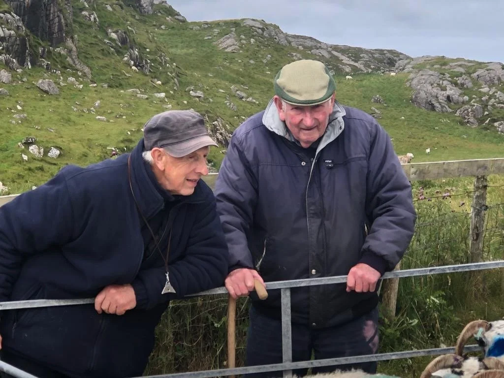 Two elderly men leaning on a metal fence, engaging with a small animal in a rural, hilly field with grass and sheep in the background.