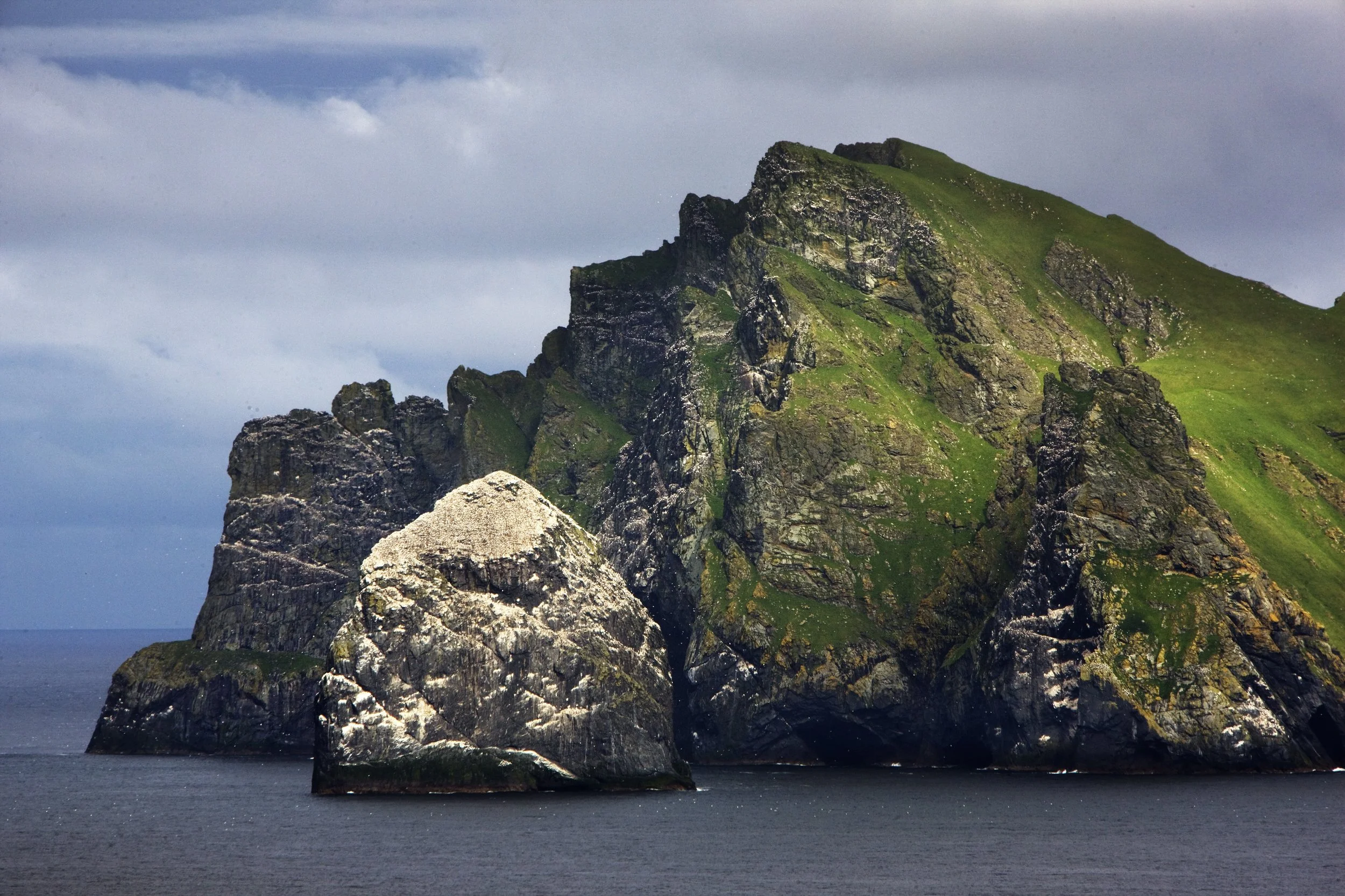 A large rocky island with steep cliffs covered in green grass, surrounded by dark water and a cloudy sky.