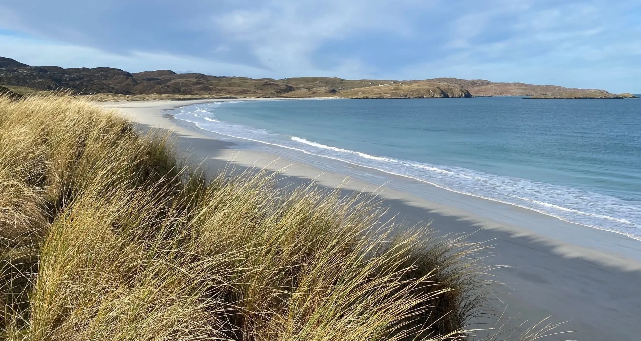 A sandy beach with tall grass in the foreground, blue ocean waves, and hills in the background under a partly cloudy sky.