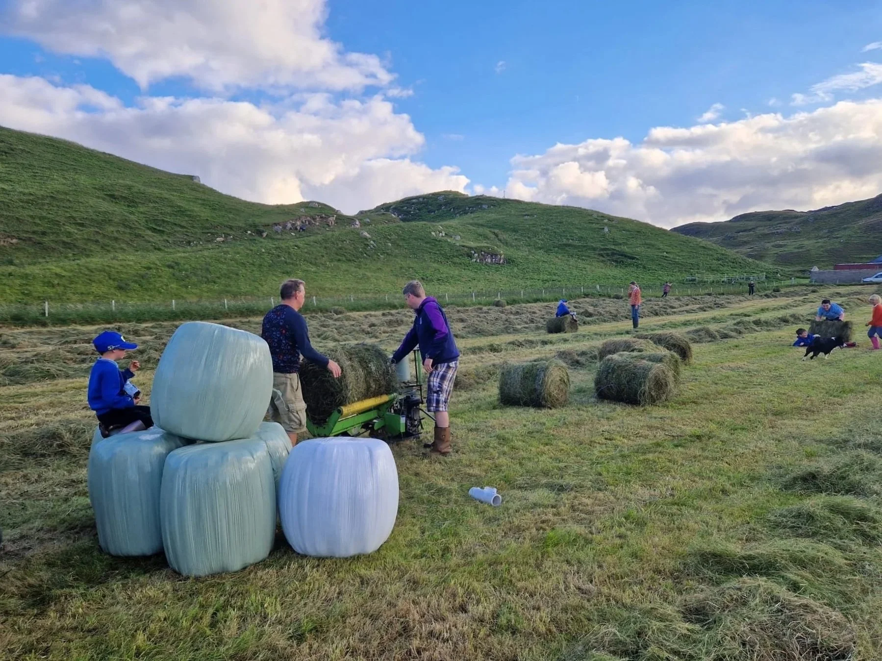 People, including children, working in a grassy field with hay bales, surrounded by green hills and a partly cloudy sky.