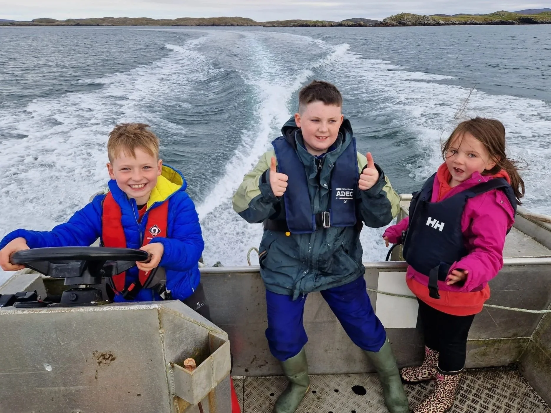 Three children on a boat, with water and land in the background. The child in the middle is giving a thumbs-up, and the children on either side are smiling or looking surprised.