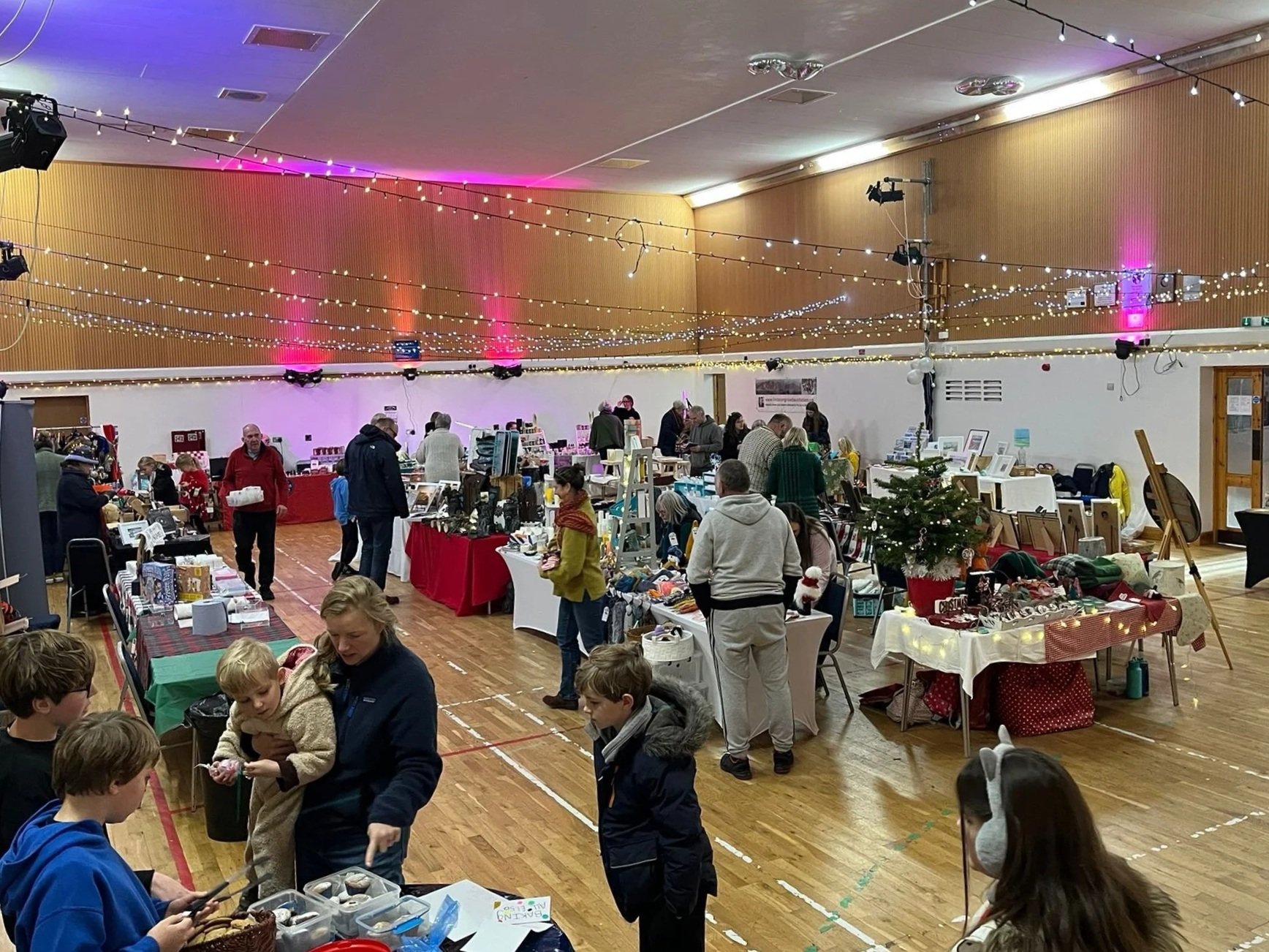 People browsing and shopping at a Christmas craft fair with decorated tables, holiday decorations, and a Christmas tree in the venue.