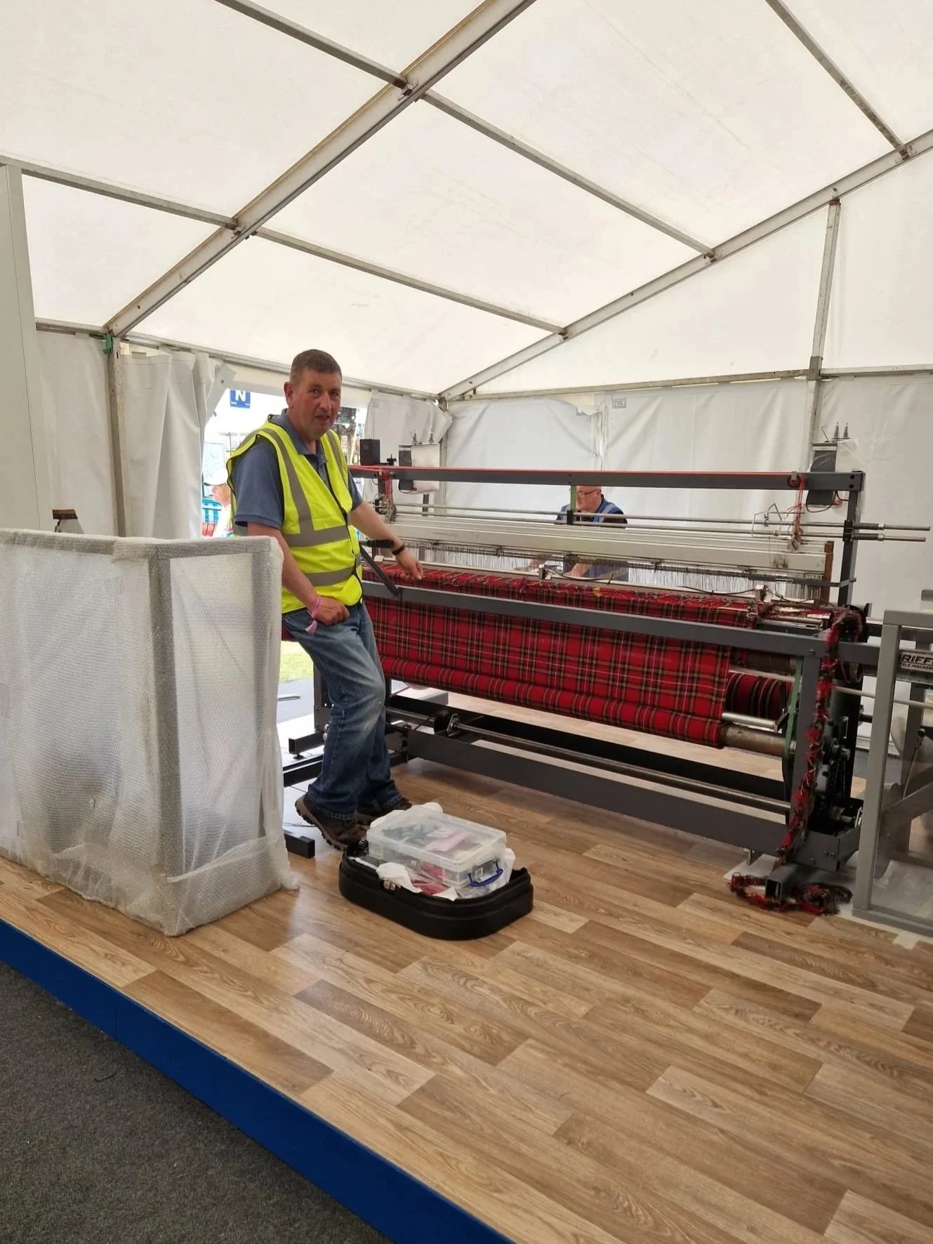 A man in a yellow safety vest standing next to a fabric quilting machine inside a white tent, with a toolbox on the floor in front of him.