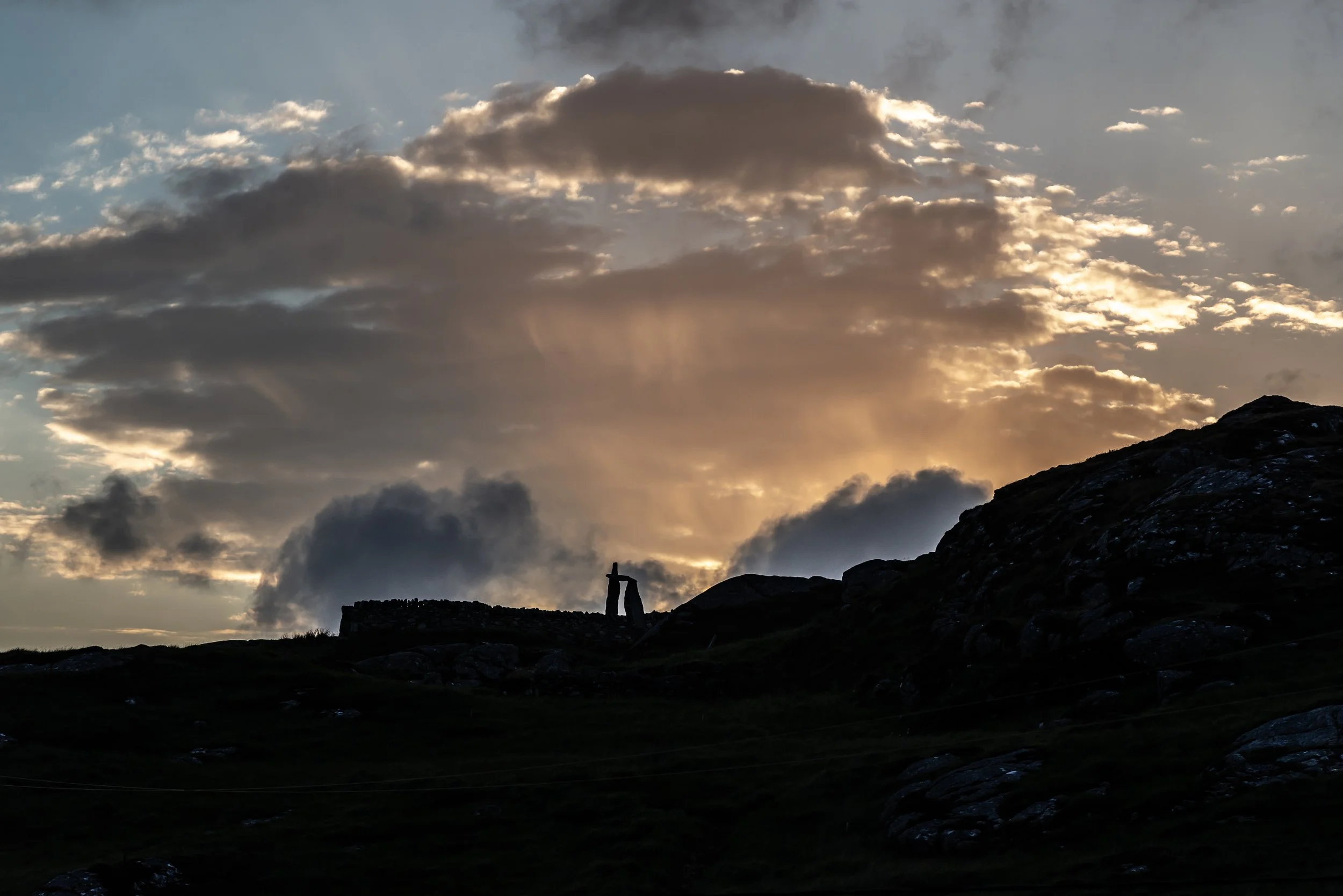 Silhouette of a landscape with a standing stone structure against a dramatic sky with clouds and sunlight. Uig, Isle of Lewis