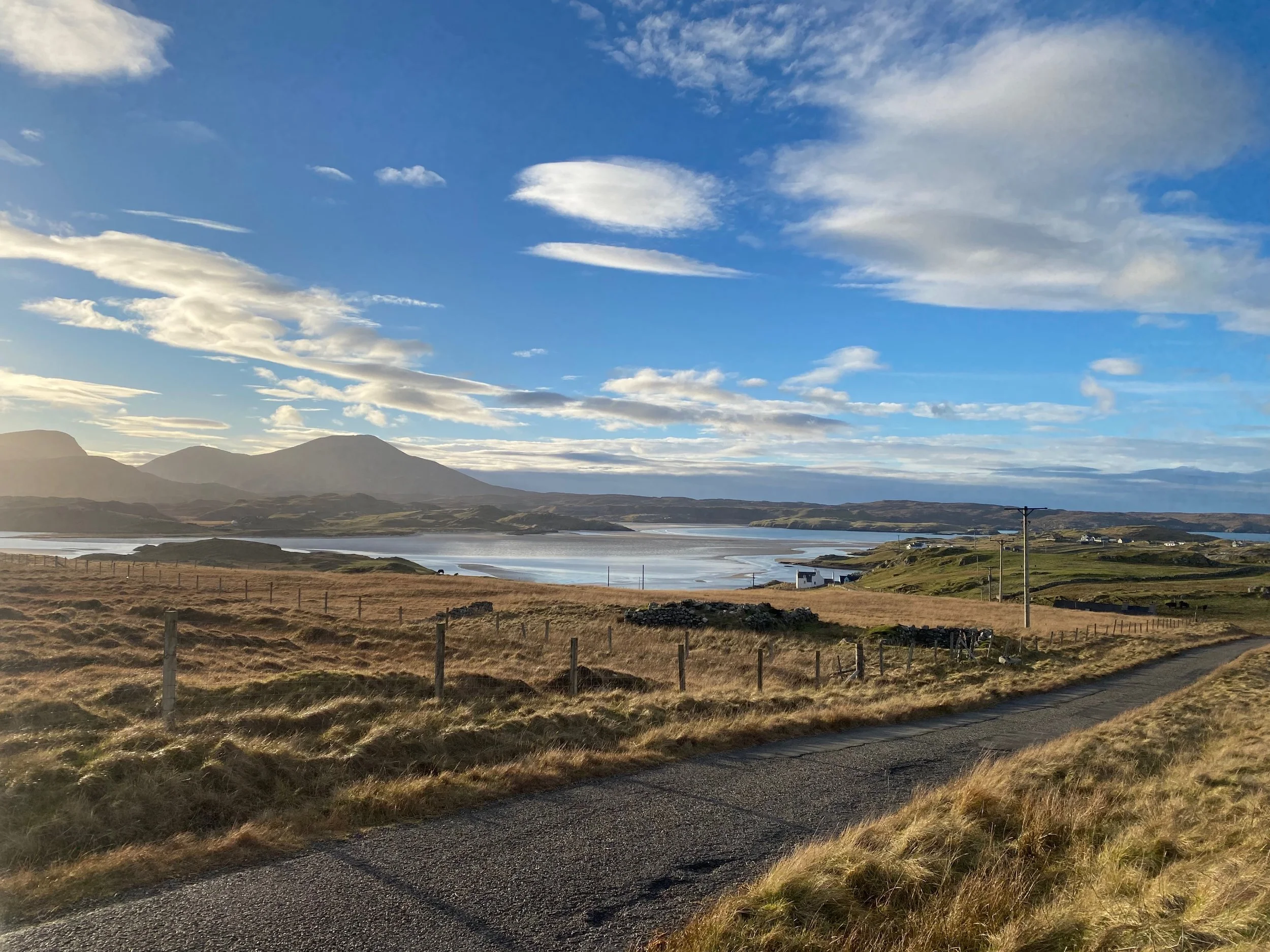 A scenic landscape of rolling grassy hills, a winding road, a body of water, mountains in the distance, and a partly cloudy blue sky with sunlight.