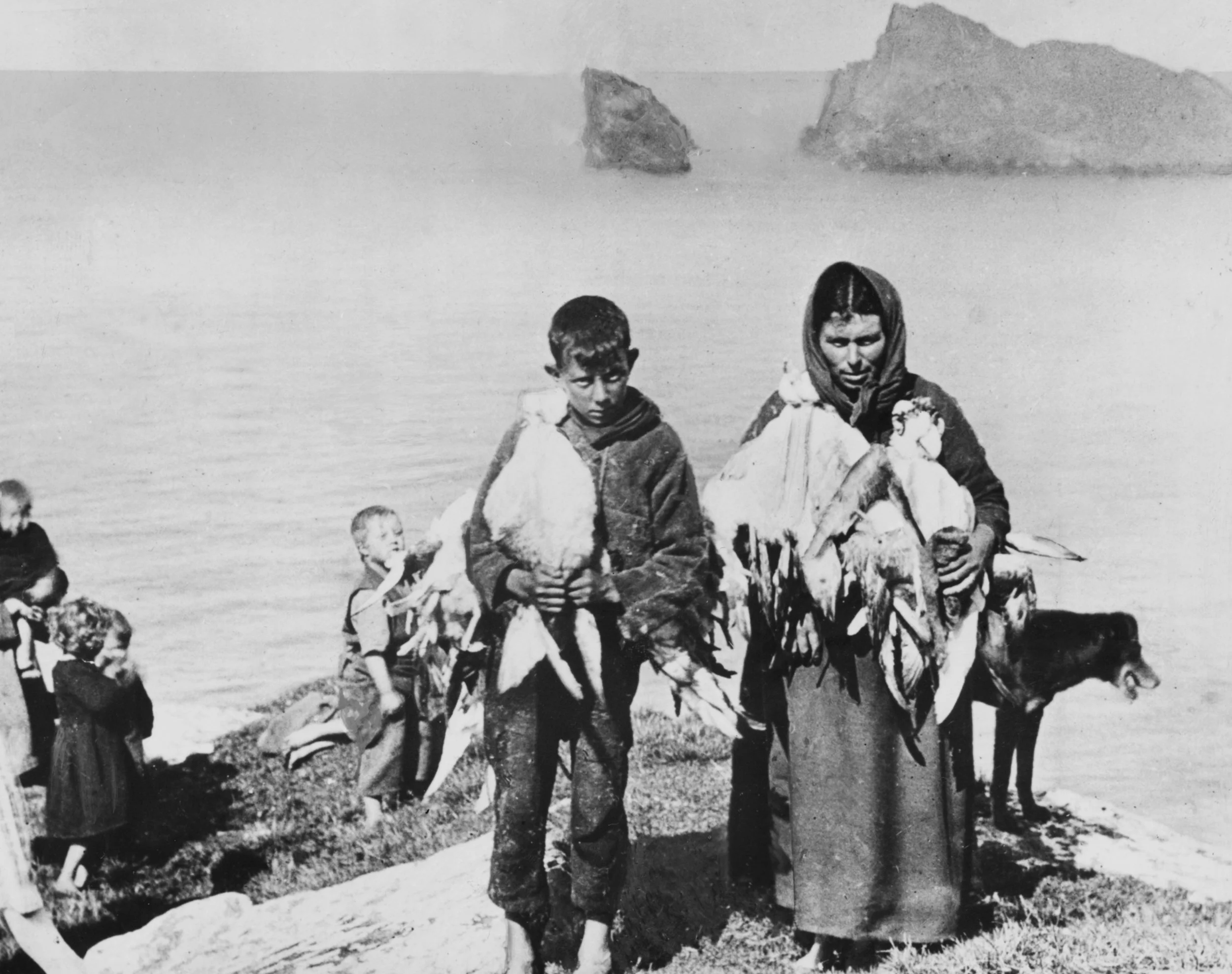 A black and white photo of a woman and a boy holding fish on a shoreline, with children in the background by the water, and rocky formations in the distance.