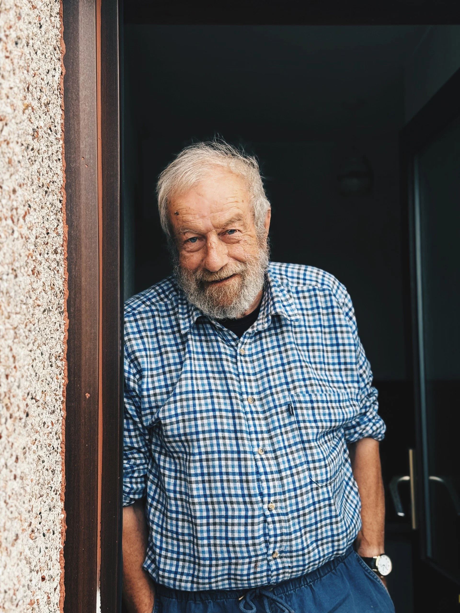 An elderly man with gray hair and beard leaning with his right shoulder against a wall, smiling while looking at the camera. He is wearing a blue checkered shirt and dark pants, with his left hand in his pocket.