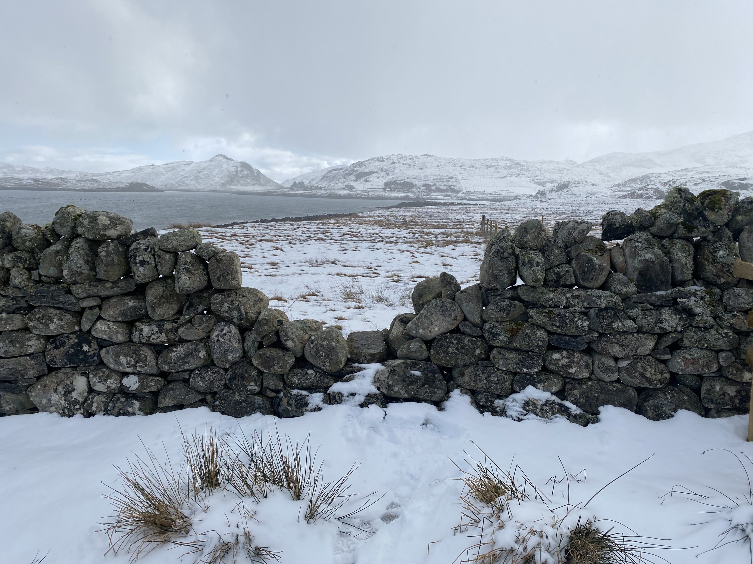 Snow-covered landscape with a stone wall in the foreground, a body of water, and snow-capped mountains in the background.