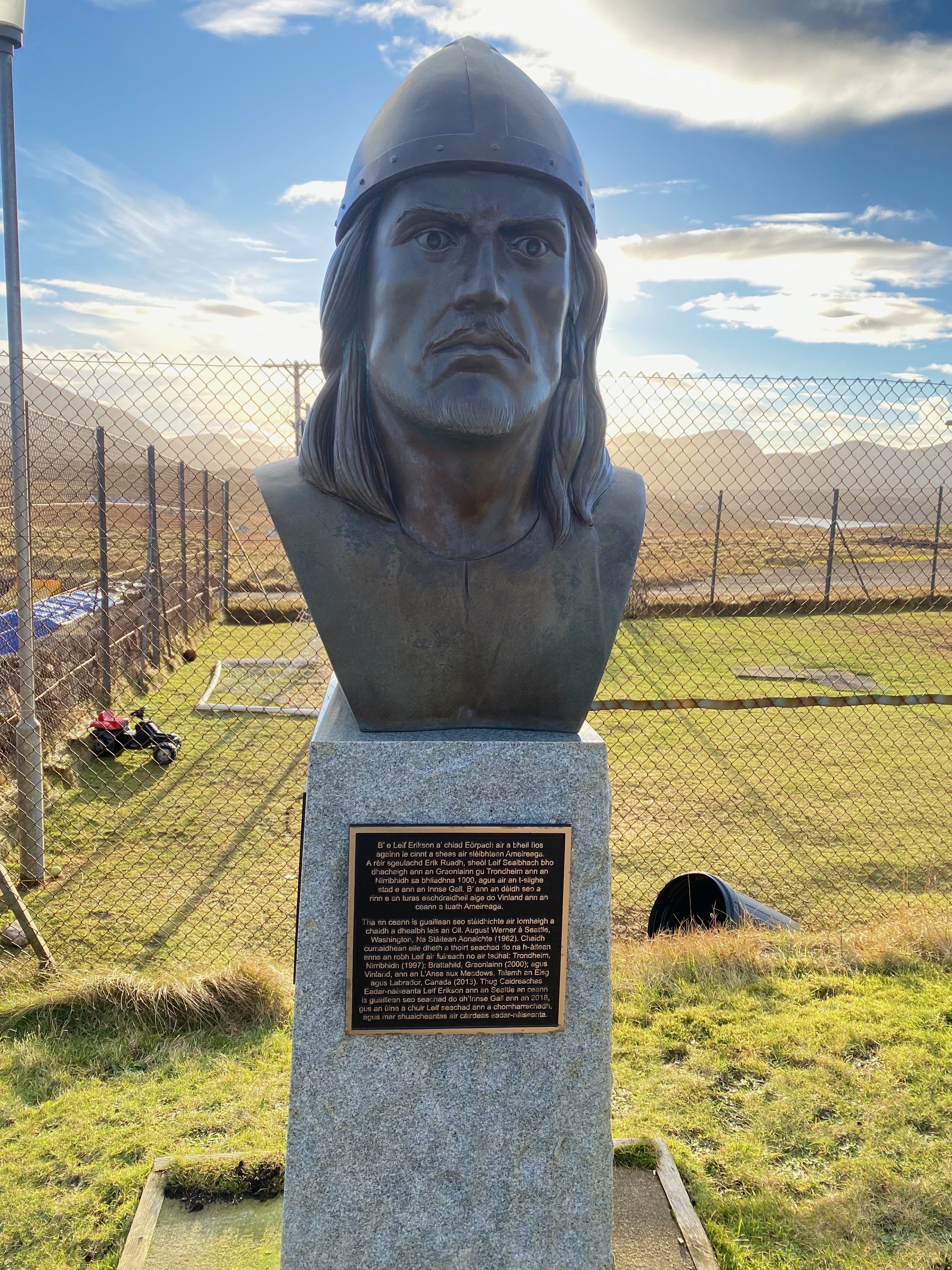 Bronze bust of a historical figure with long hair and a beard wearing a helmet, mounted on a stone pedestal with an informational plaque, outdoors with a chain-link fence and mountains in the background.