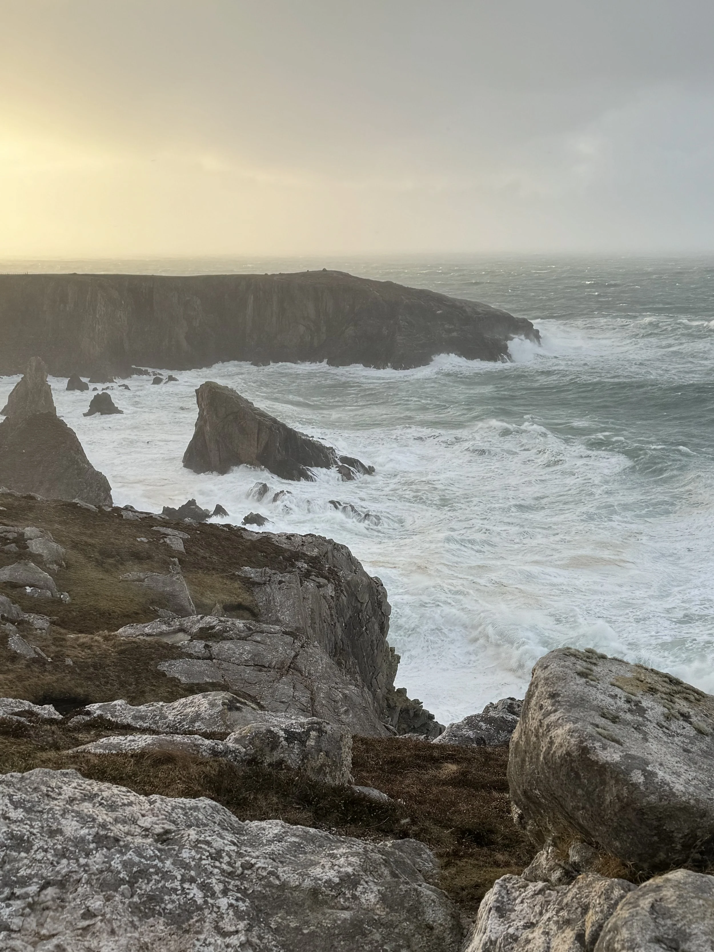 A rugged coastline with large rocks and crashing waves, under a cloudy sky with a hint of sunlight.
