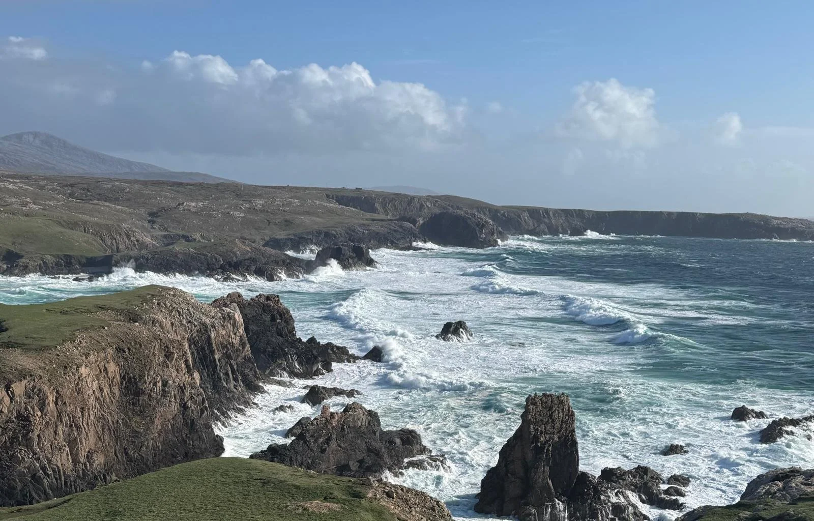 Coastal landscape with rocky cliffs, rolling waves, and grassy terrain under a partly cloudy sky.