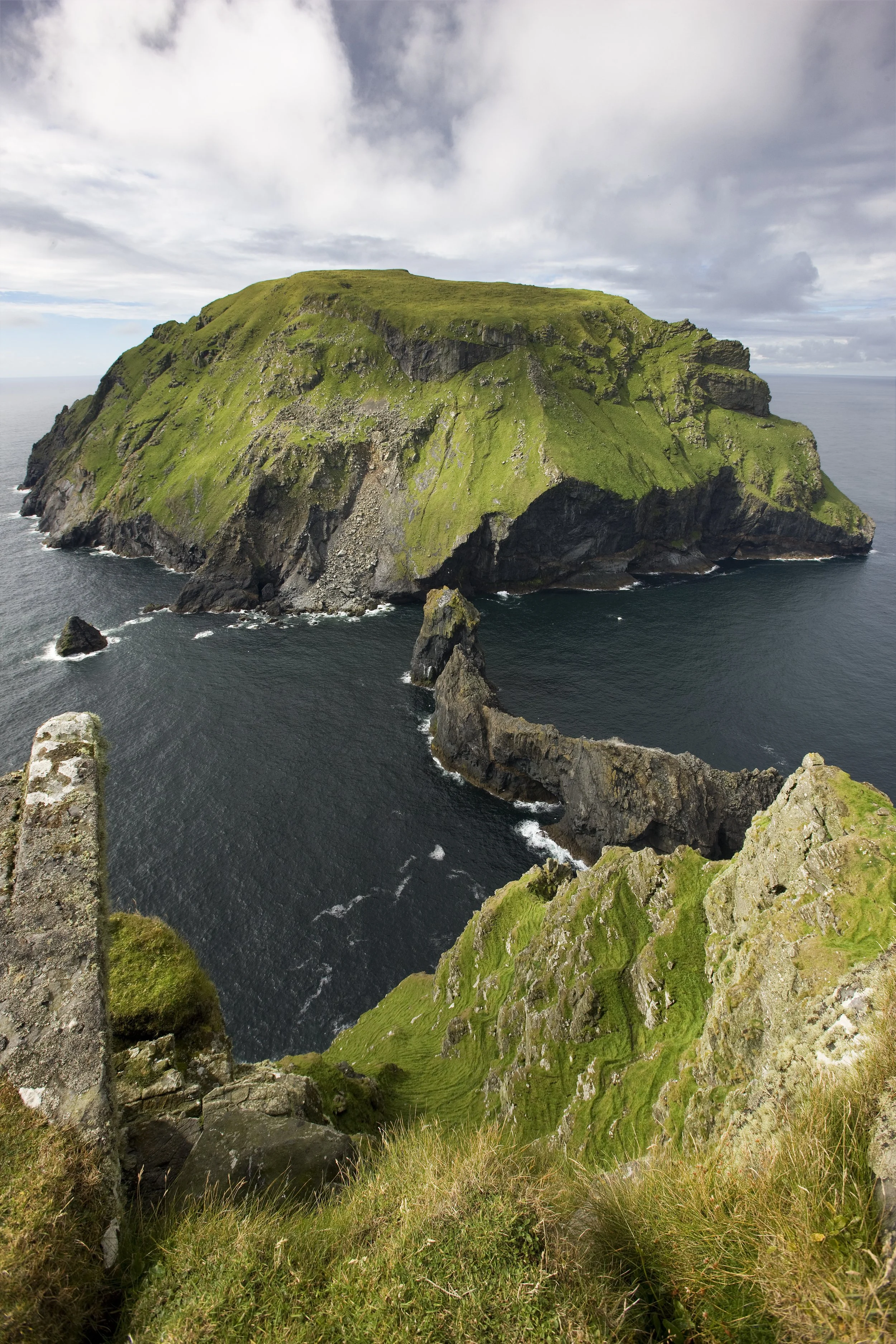 A large rocky island covered in green grass surrounded by ocean water, with a cloudy sky overhead.