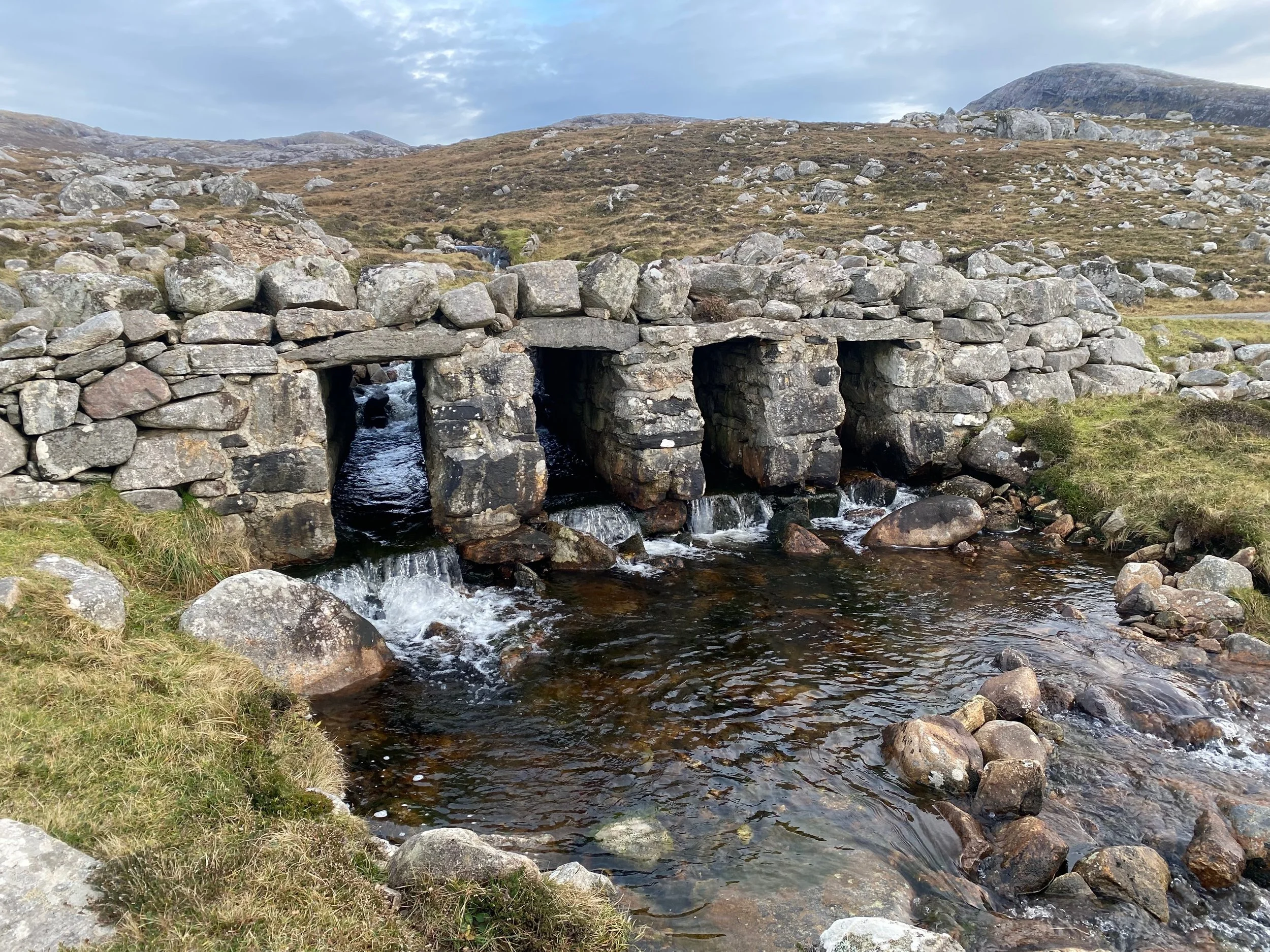 A small stone bridge with three arched openings over a flowing stream in a hilly landscape with rocky terrain and sparse grass.