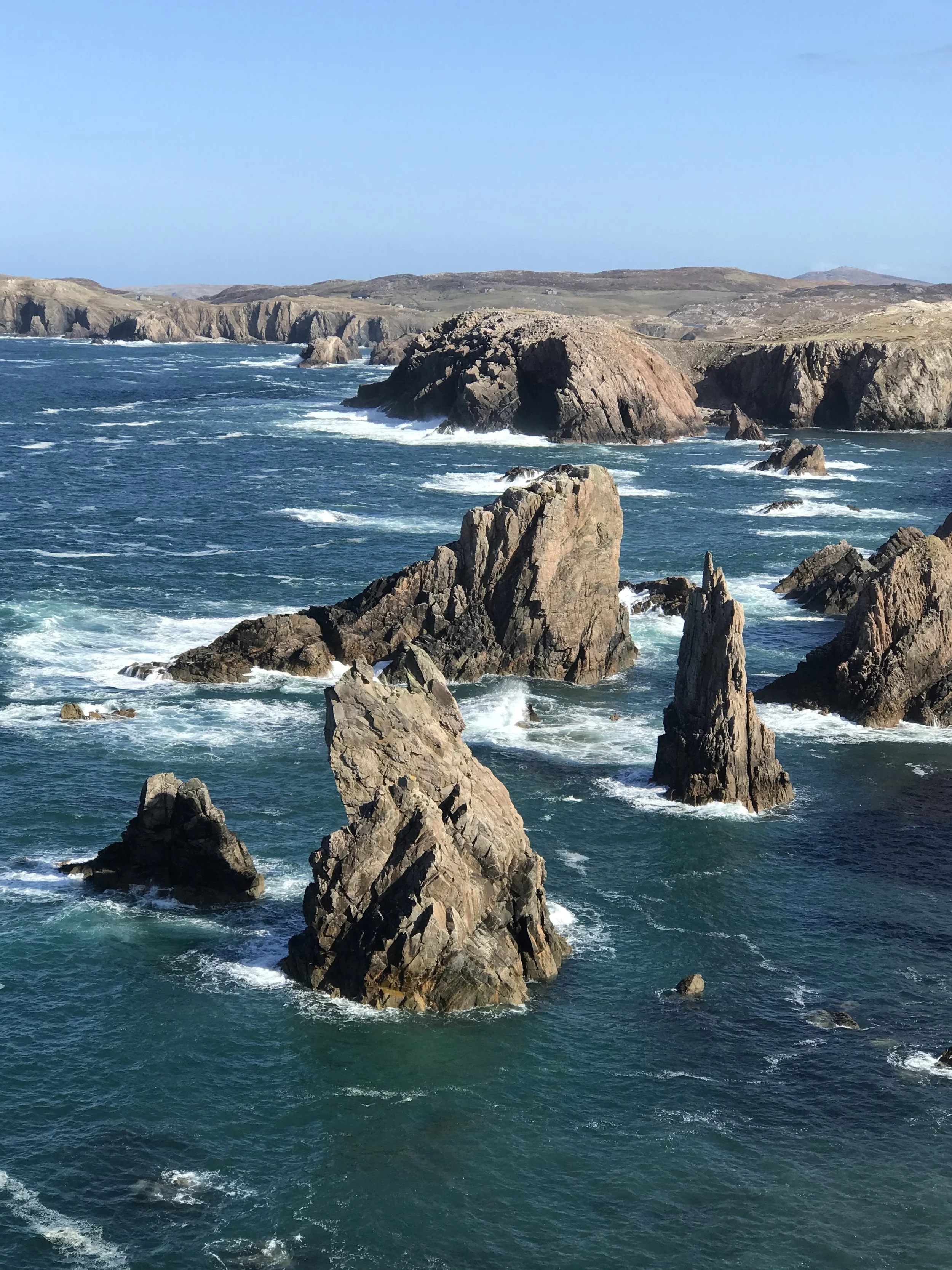 Rocky coastline with tall jagged rocks protruding from the ocean, waves crashing against the rocks, and a distant view of cliffs under a blue sky.