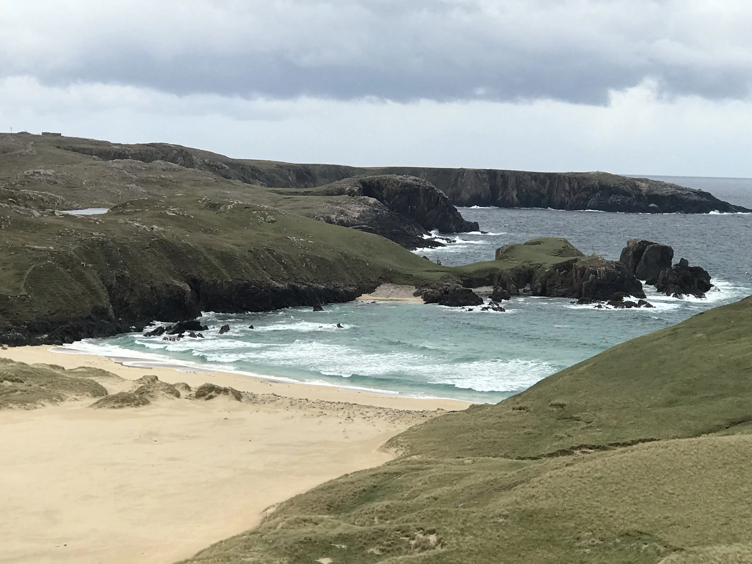Overcast day at a secluded beach with white sand, rocky cliffs and green grassy hills surrounding the shoreline, with the ocean on the right.