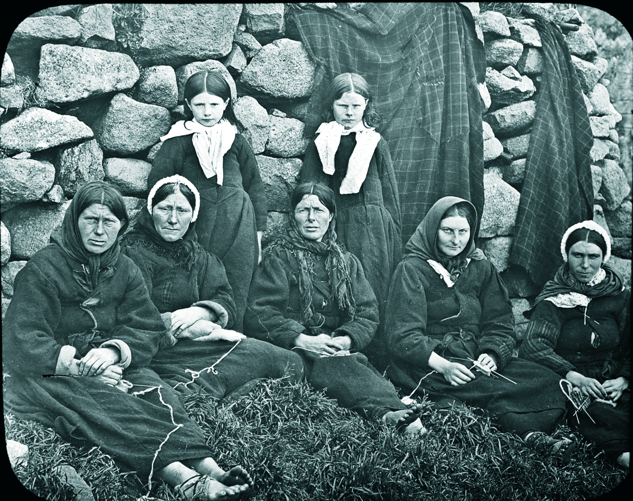 Seven women and two girls sitting and standing in front of a stone wall. The women are dressed in traditional, modest clothing with headscarves, and some appear to be knitting or holding yarn. The girls are also dressed modestly, with one girl standing and the other sitting. It appears to be an old black-and-white photograph.
