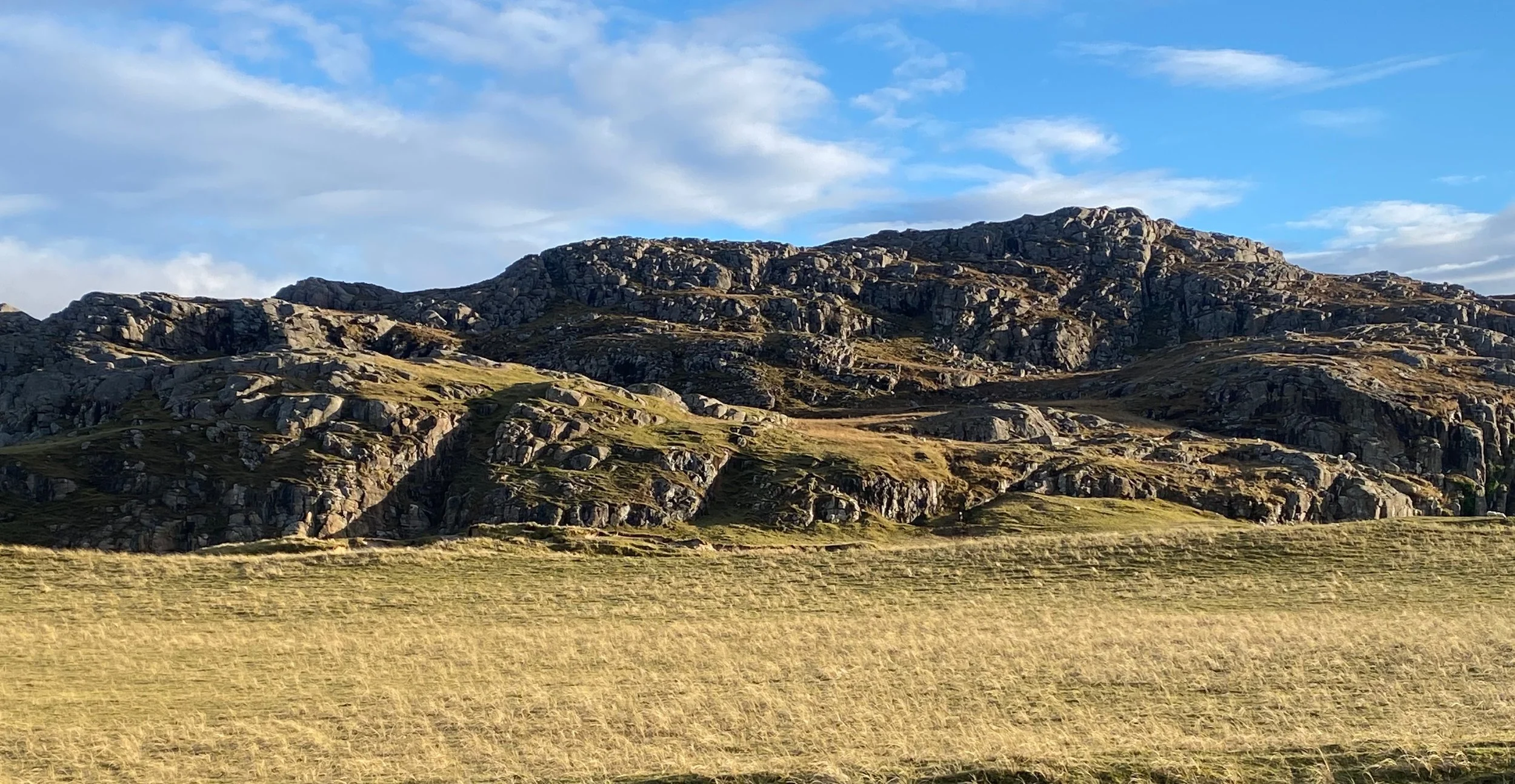 A rocky mountain with sparse vegetation under a partly cloudy sky, with a grassy plain in the foreground.
