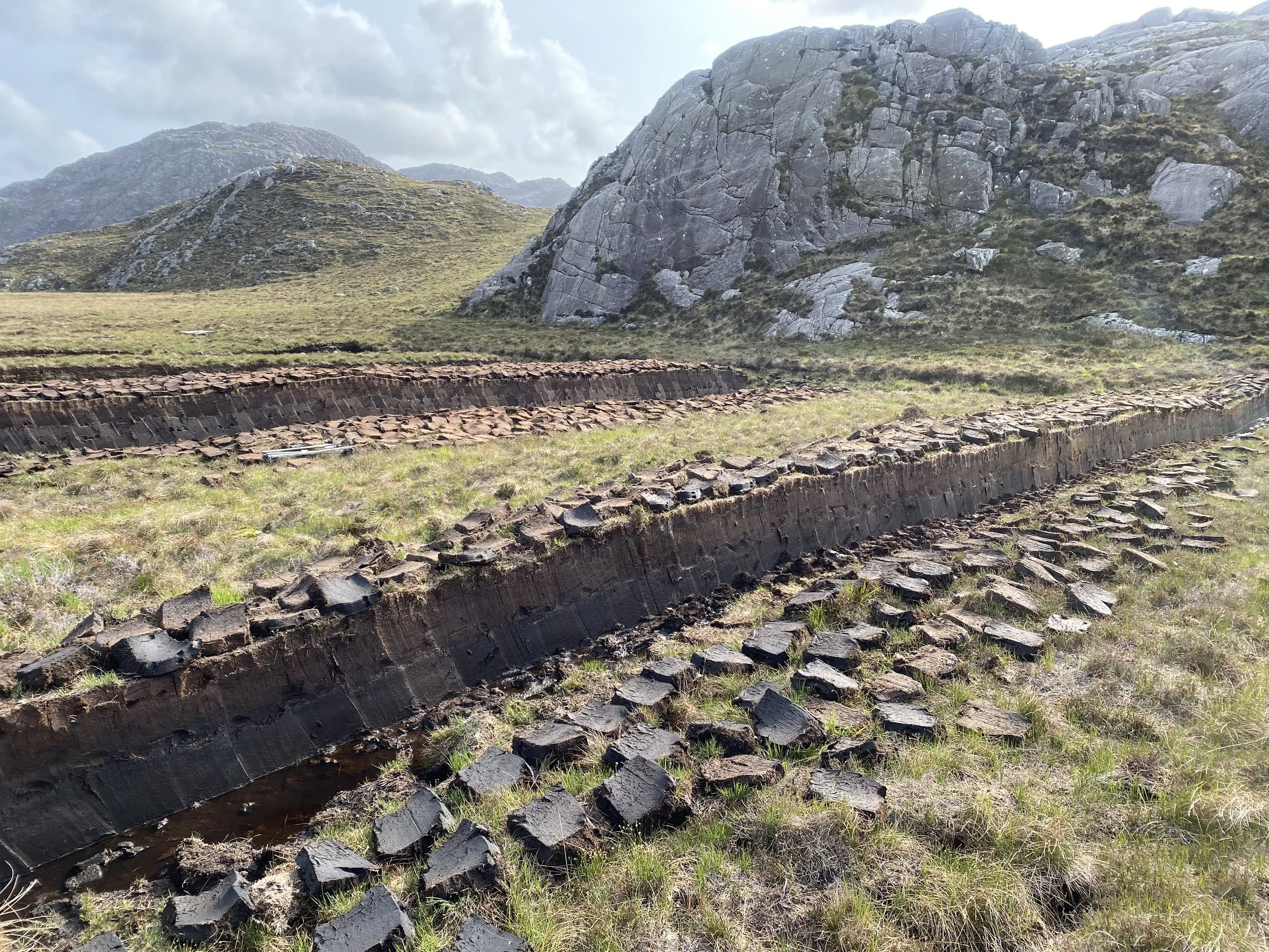 Ruins of an ancient stone structure in a grassy landscape surrounded by mountains under cloudy sky.