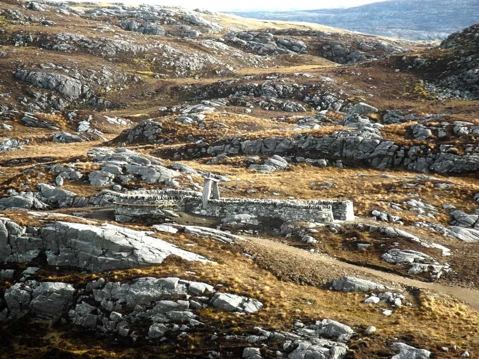 Rugged mountain landscape with rocky slopes and patches of grass, featuring an old stone structure or wall and a small cross-like monument.