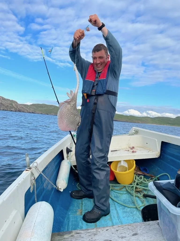 A man in a blue and red life jacket, standing on a boat, holding a fishing rod with a large fish caught on the line. He is smiling, with a scenic water and hilly landscape in the background.