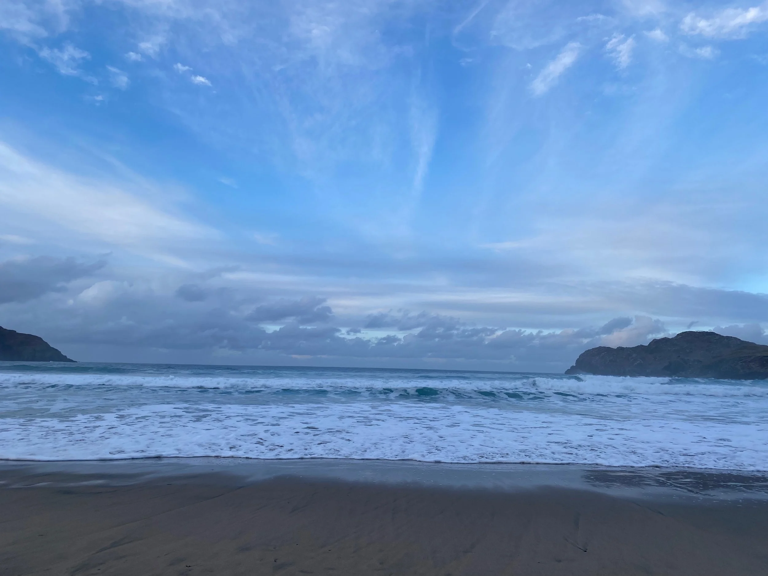 A scenic view of the ocean with waves crashing on the sandy beach, and rocky hills in the distance under a partly cloudy blue sky.