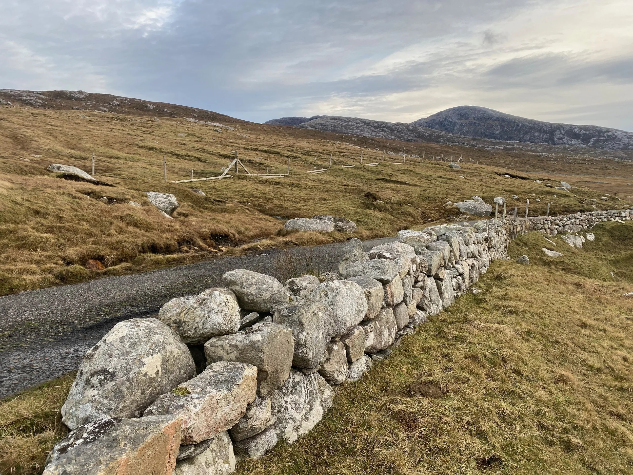 A stone wall beside a paved pathway in a grassy hilly landscape with mountains in the distance and a cloudy sky.