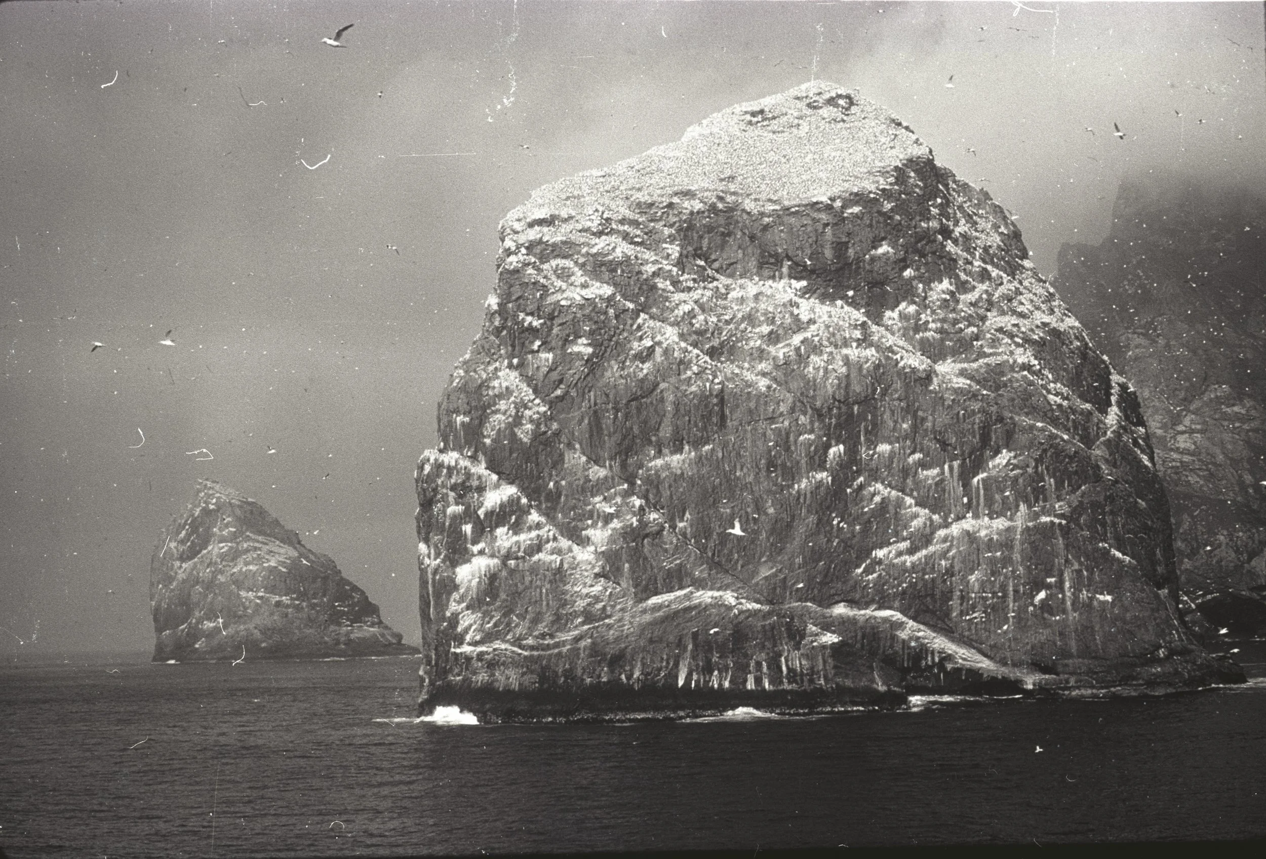Black and white photograph of a large rocky sea stack with a smaller rock formation in the background, surrounded by water and seagulls flying overhead.