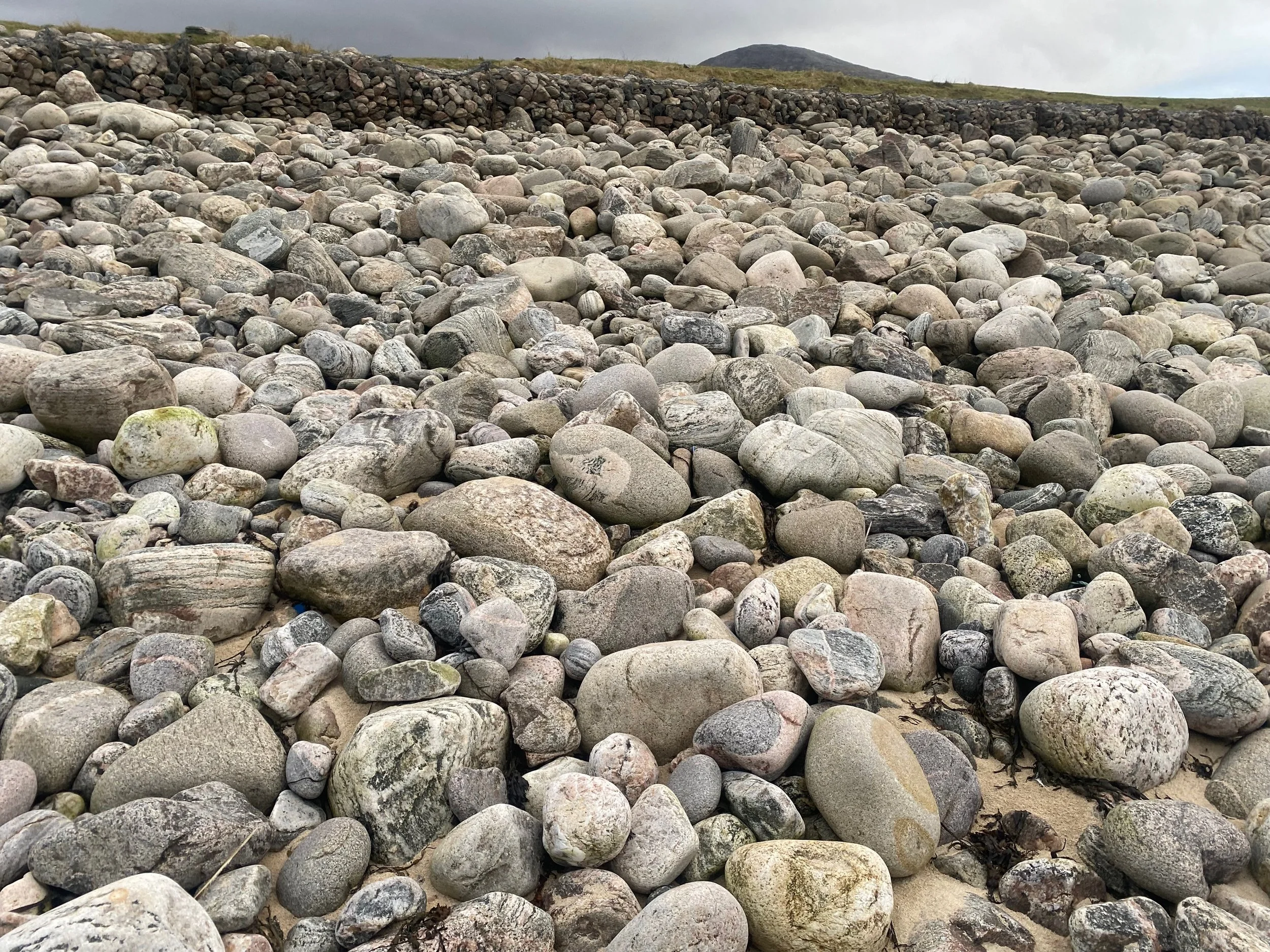 A rocky beach with numerous smooth, rounded stones of various sizes and shades of gray, with a stone wall in the background and a cloudy sky.