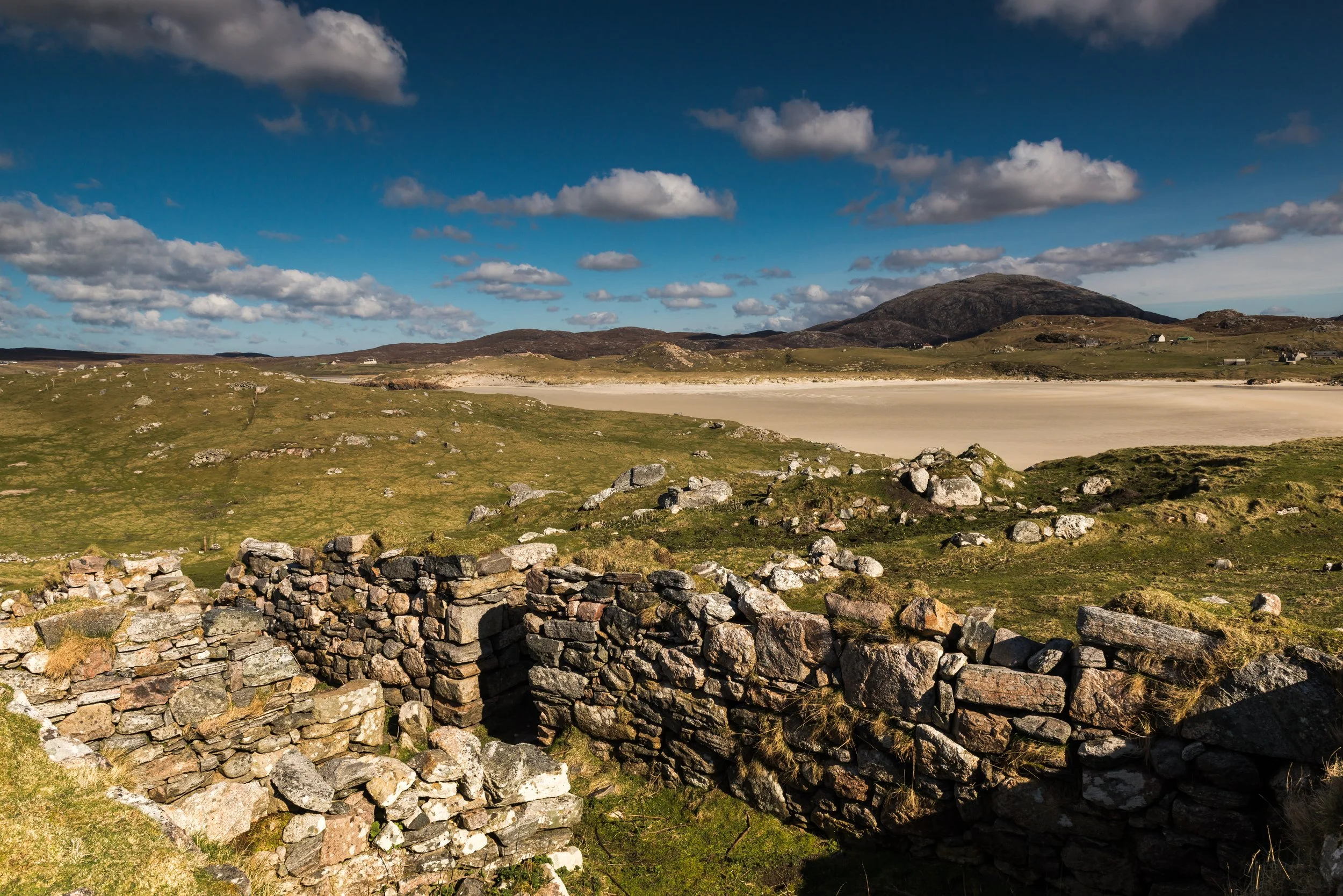Scenic landscape with stone ruins, grassy fields, a sandy beach, hills, and a mountain under a partly cloudy sky. Uig, Isle of Lewis