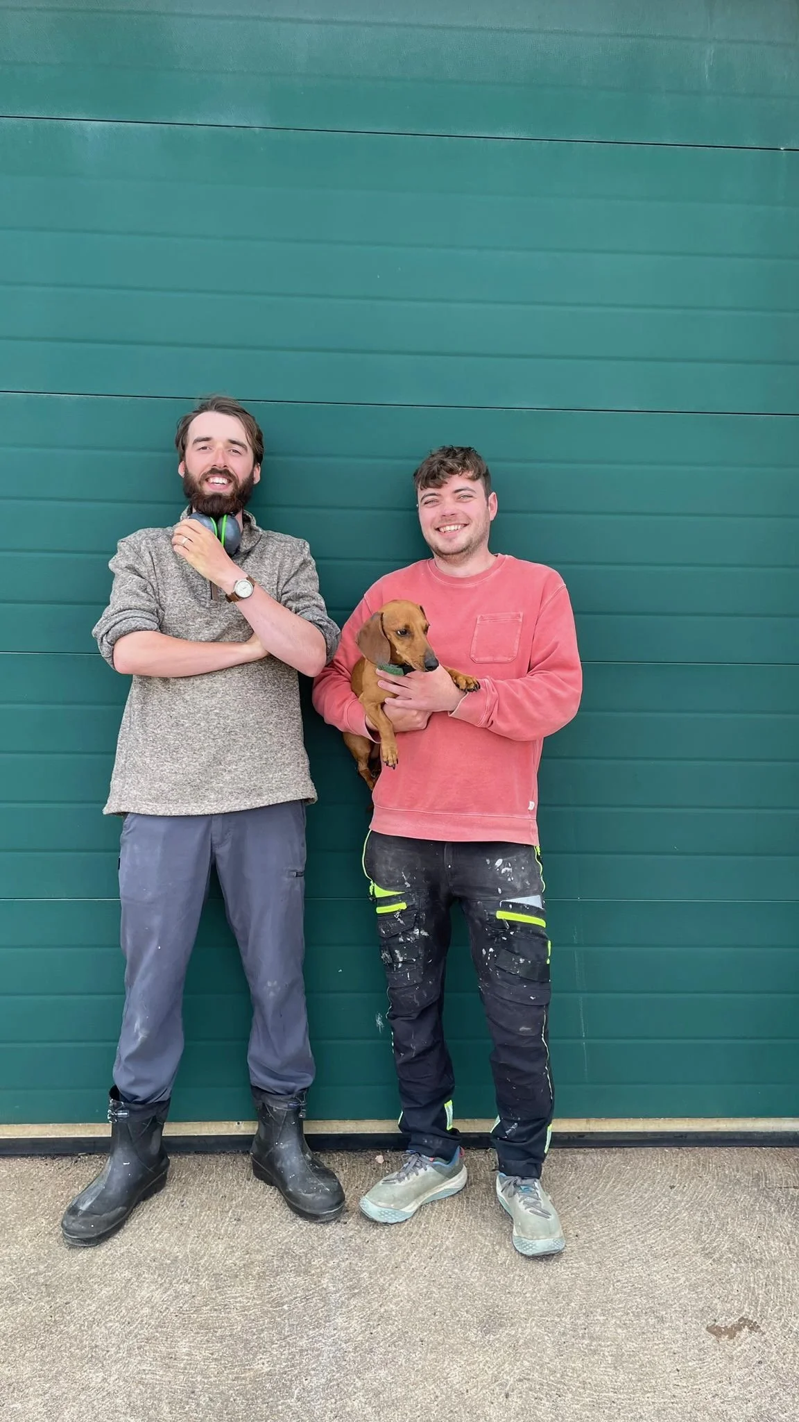 Two young men standing in front of a green garage door, smiling; the man on the right is holding a brown Dachshund puppy.