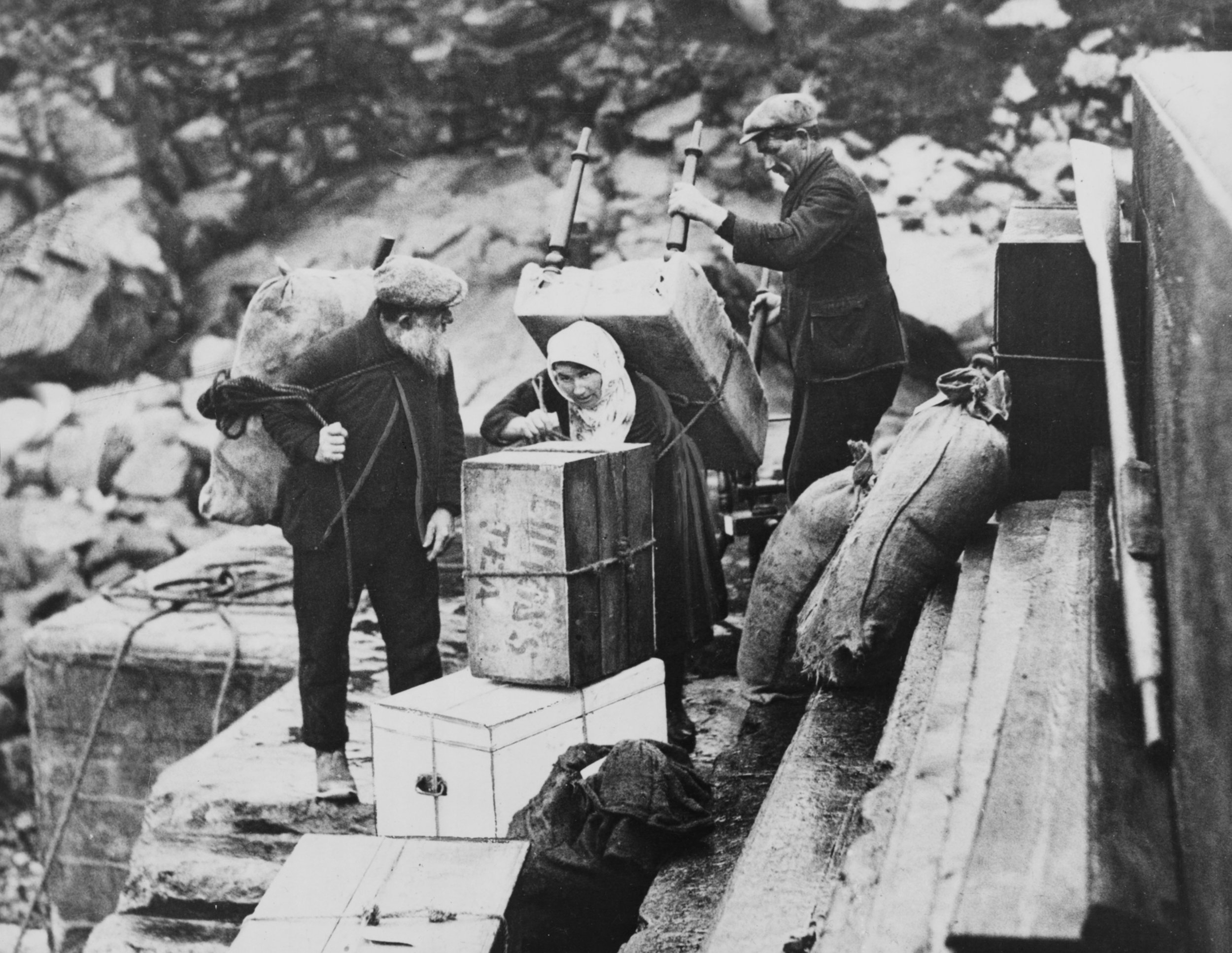 Three men and one woman standing on a wooden platform by a river, with two of the men holding large guns pointed downward, and the woman looking into a box or container, surrounded by boxes and bags.