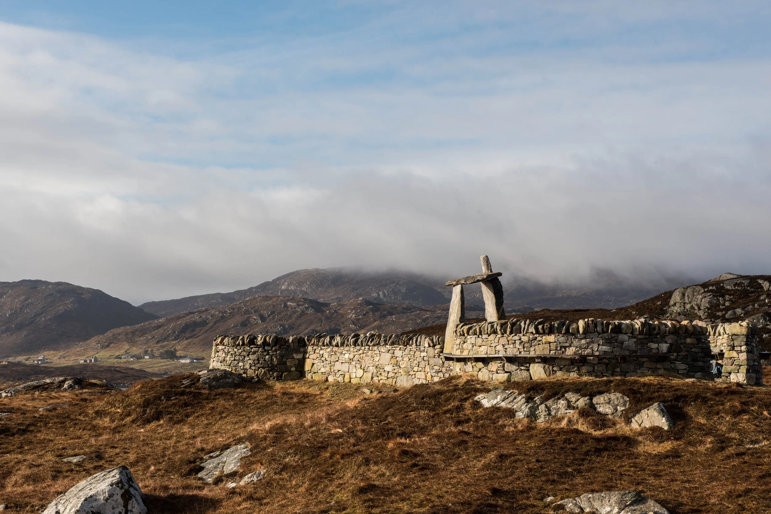 Stone wall and a simple stone sculpture in a mountain landscape with cloudy sky and distant hills.