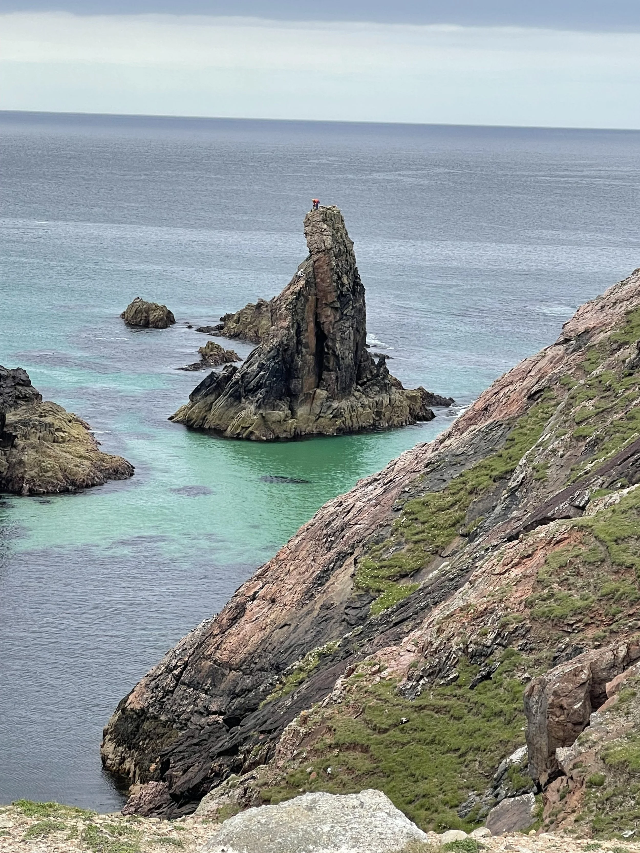 Rocky coastal landscape with a tall, jagged rock formation protruding from the turquoise water, surrounded by smaller rocks and green moss-covered cliffs under an overcast sky.