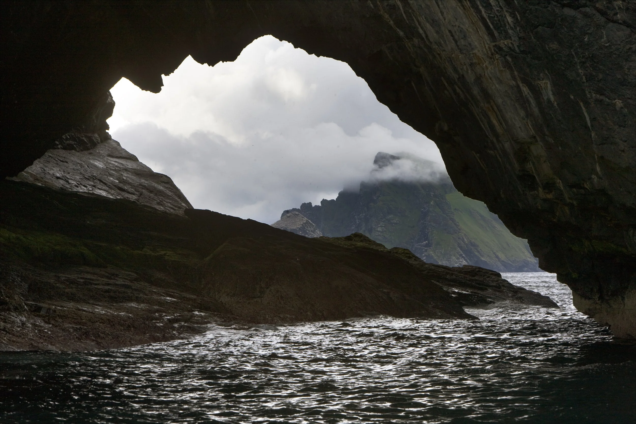 View from inside a sea cave looking out to rocky landscape and mountains with clouds.