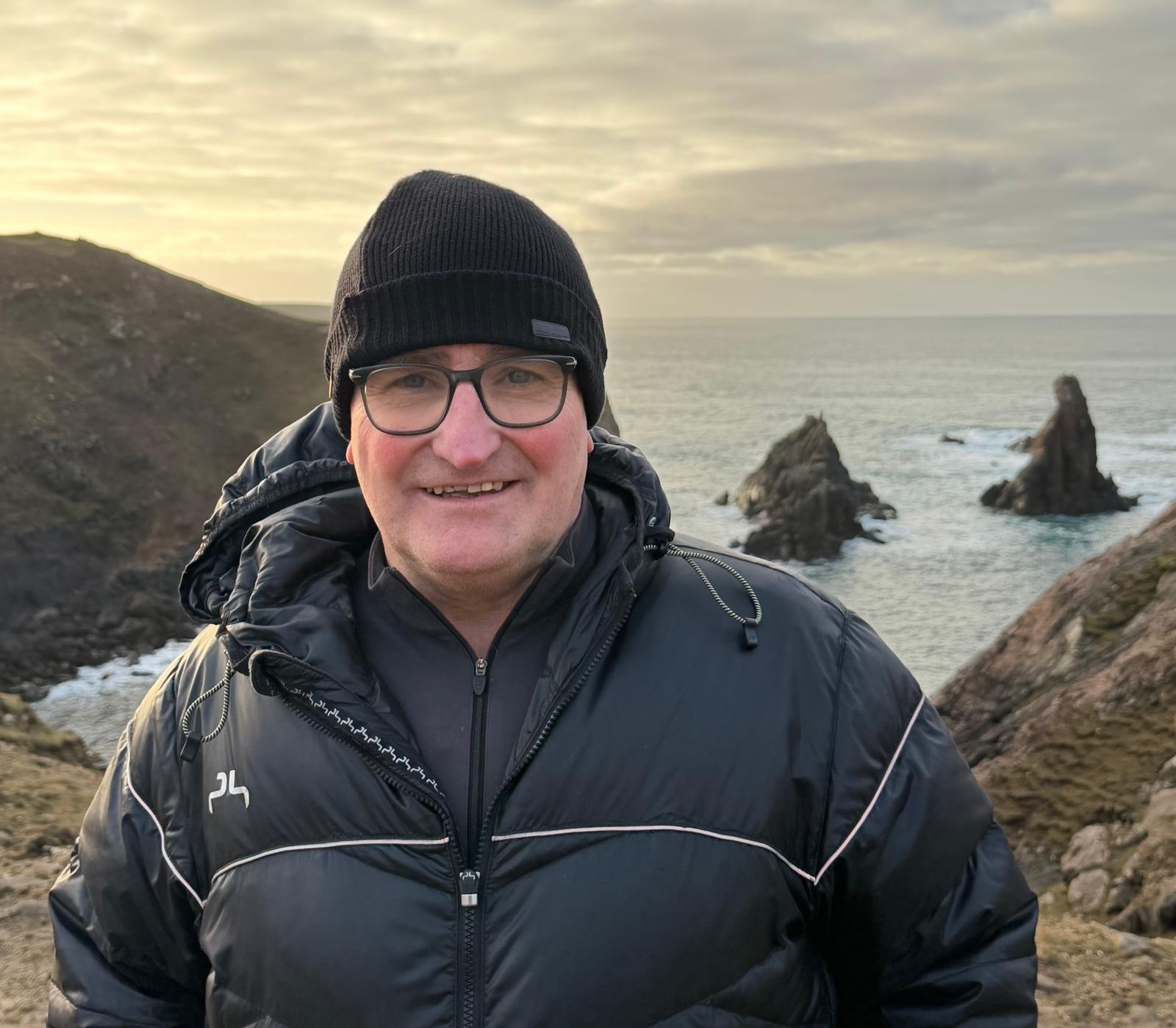 A rugged volcanic landscape with steaming vents, brownish-orange rocks, and patches of snow, under a cloudy sky.