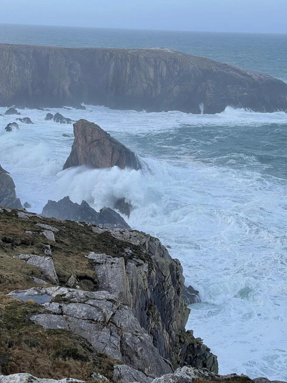 Rough ocean waves crashing against jagged rocks along a cliff coast, with large cliffs in the background under a cloudy sky.