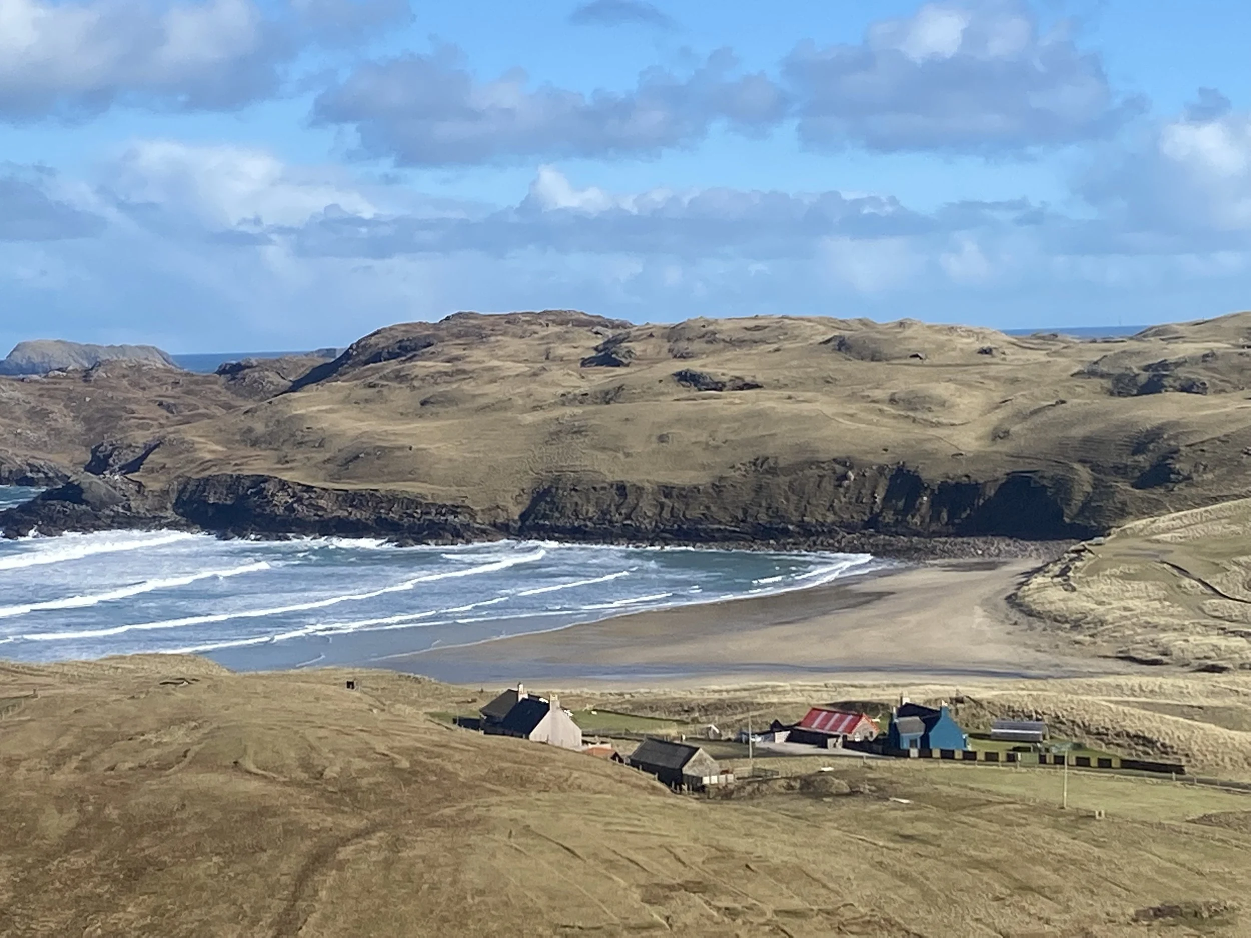 A coastal landscape featuring a beach, rolling hills, and small houses with colorful roofs under a partly cloudy sky.