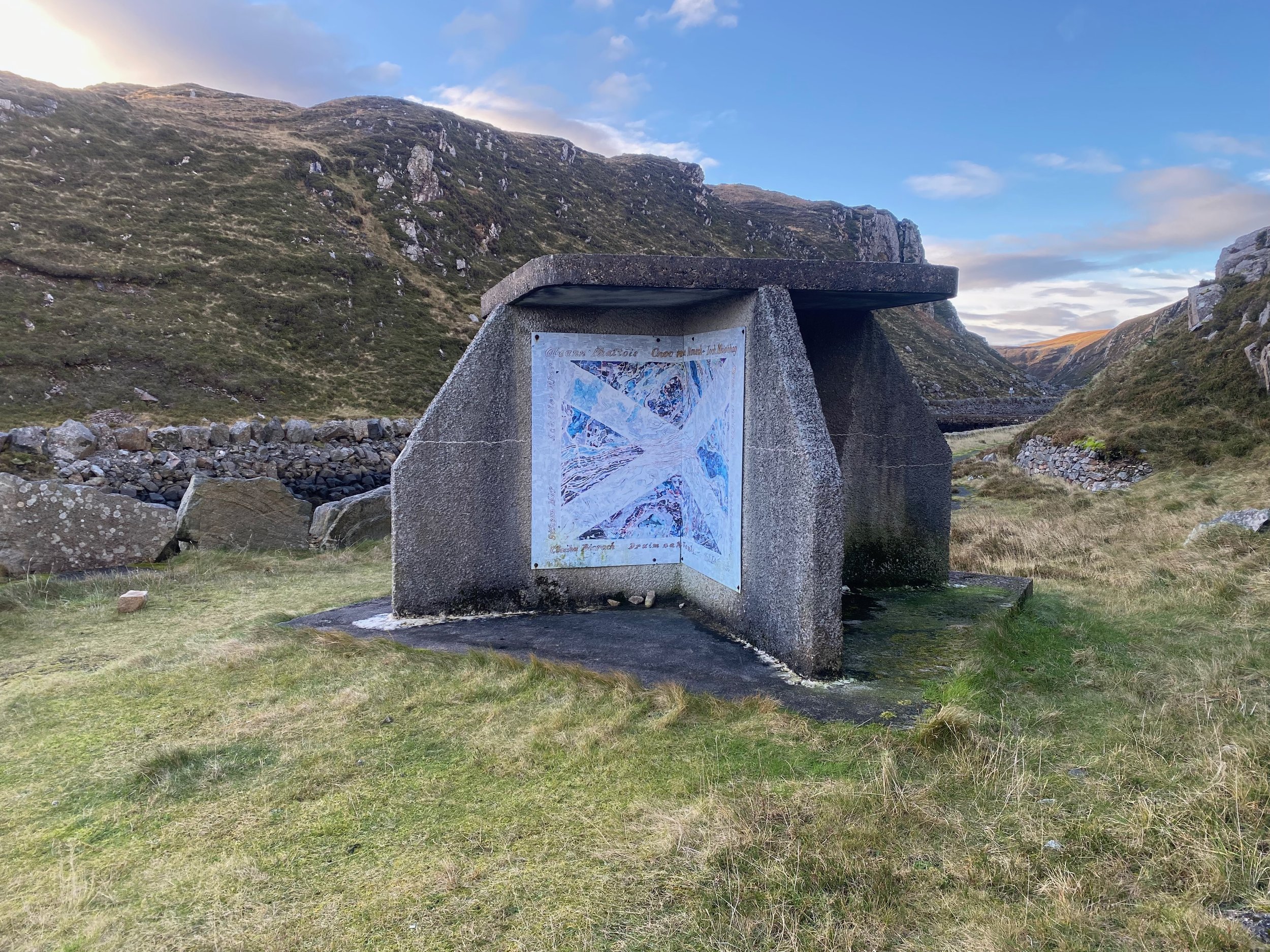 A stone memorial with a mural of the Union Jack flag in a rural, mountainous landscape with rolling hills and a cloudy sky.