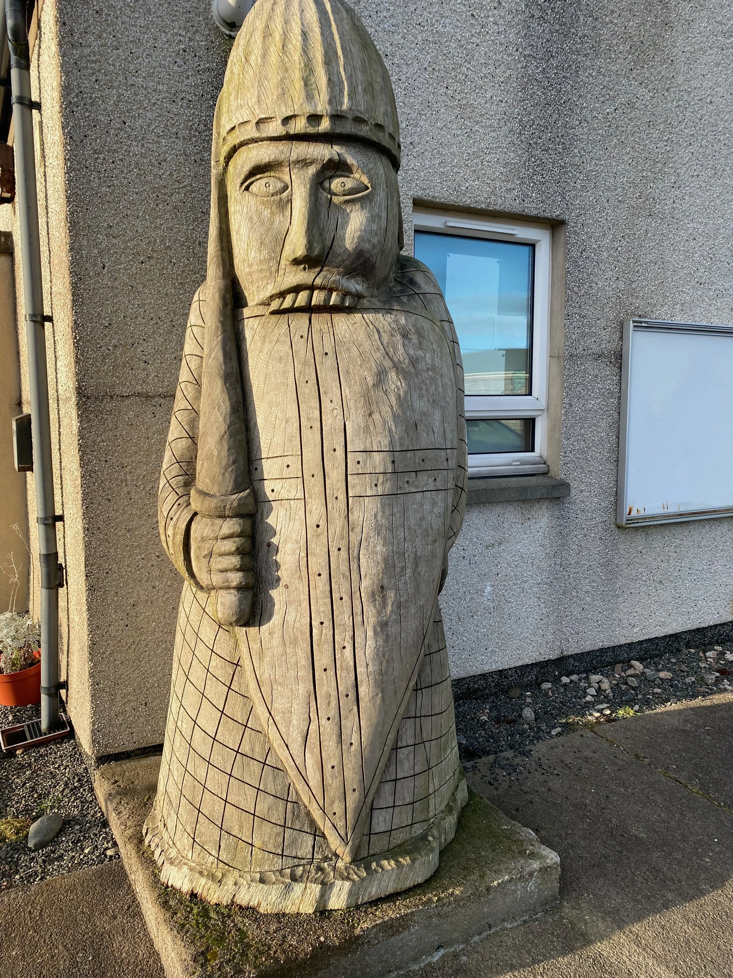 Wooden statue of a man wearing a helmet and holding a sword, standing outside next to a building with a window and a whiteboard.