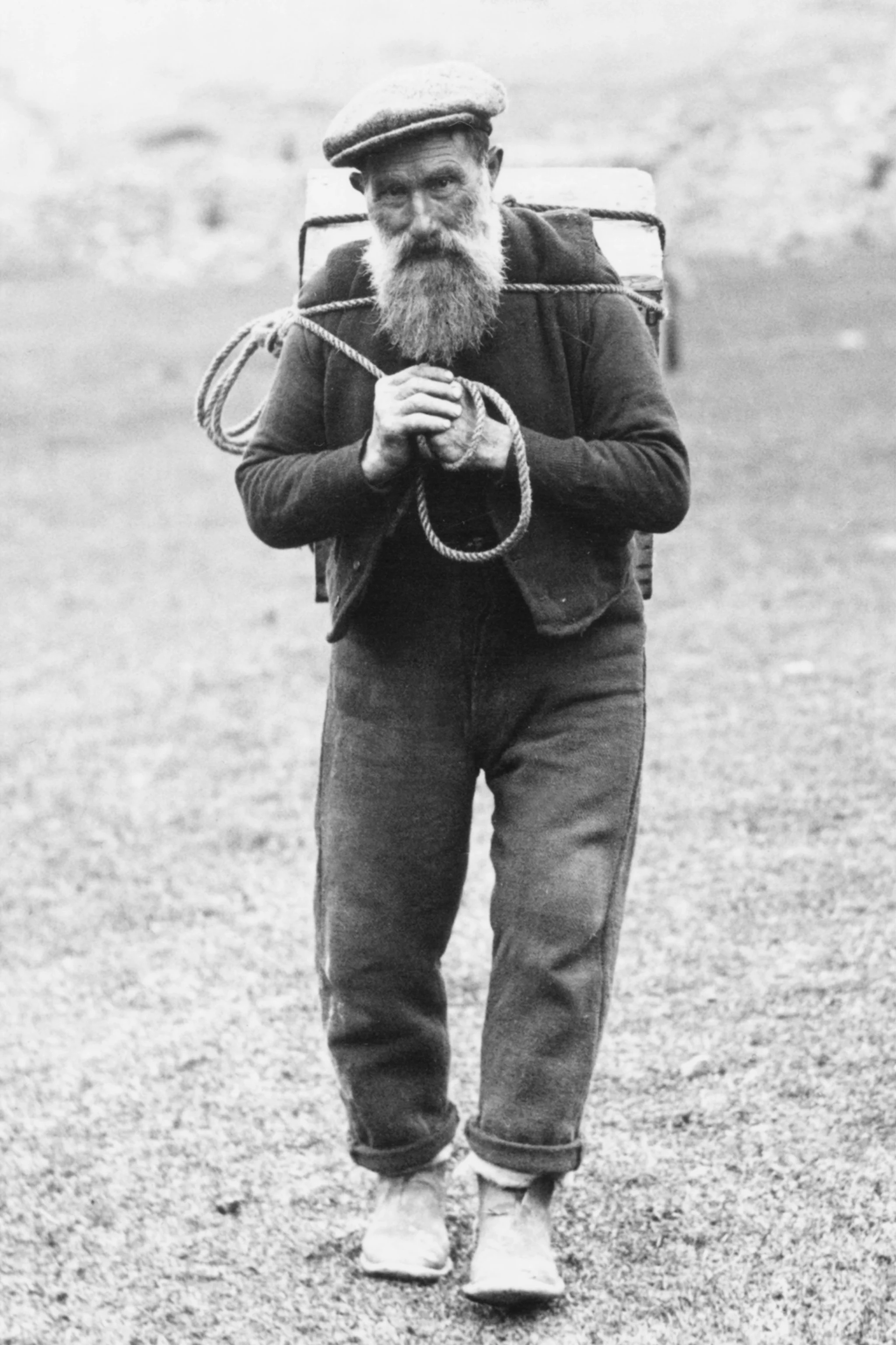 Black-and-white photo of an elderly man with a beard, wearing a flat cap, dark jacket, and rolled-up pants, carrying a backpack and holding a rope, walking on a grassy field.